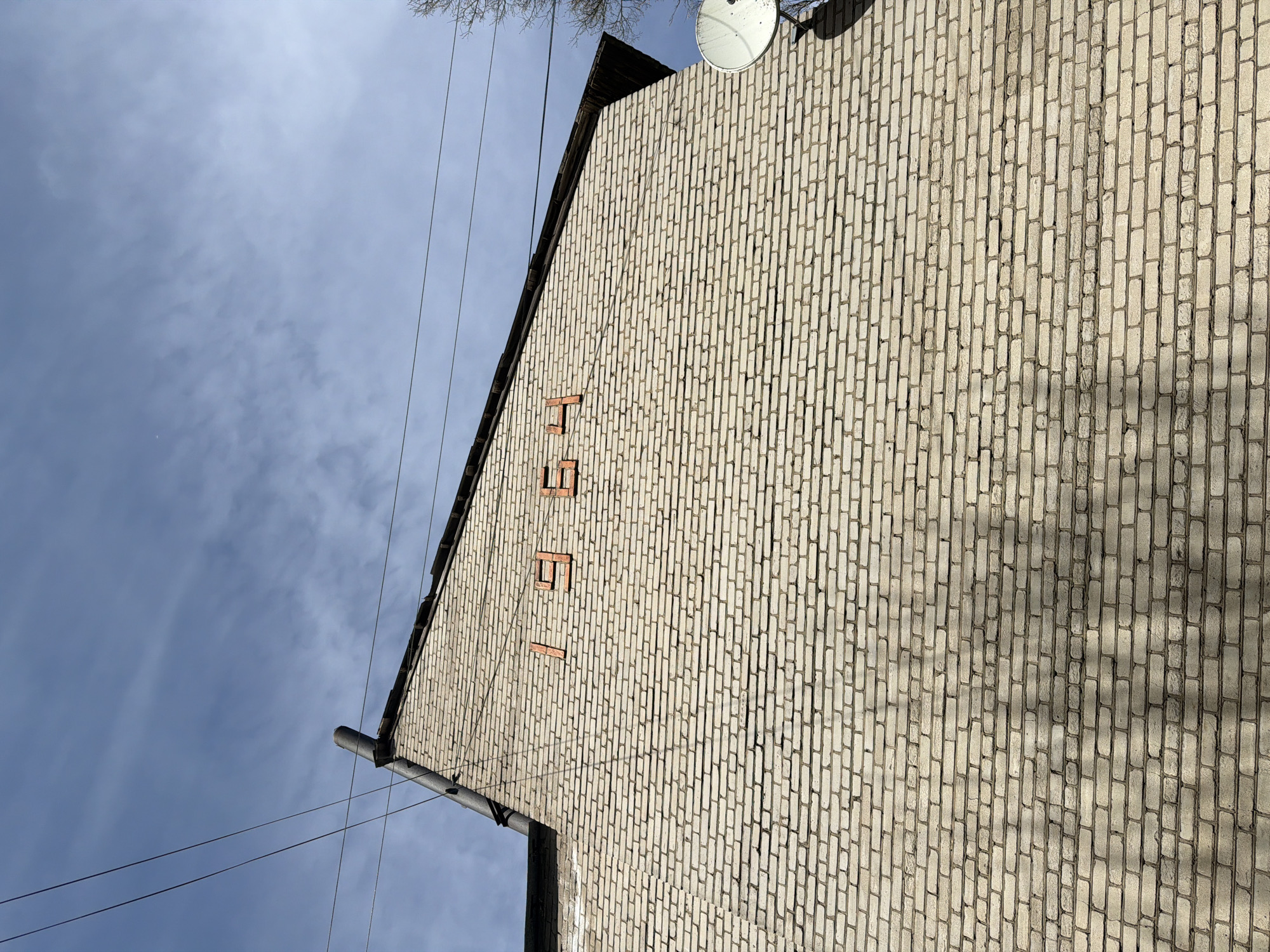 The image shows a section of a light-colored brick building in Tokkari, Russia. The facade features the numbers "167" formed by protruding red bricks. A satellite dish is mounted on the building's roof. Power lines run across the upper left of the frame against a blue sky with some cloud cover. A downspout is visible near the lower left corner of the building.