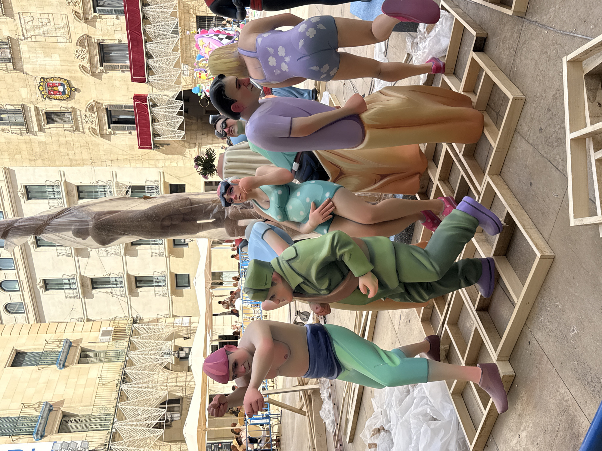 The image shows a street festival scene in an urban environment. In the foreground, several large, colorful figures are displayed on a paved plaza. These figures appear to be part of a parade or exhibition, with some dressed in exaggerated costumes. One figure wears a pink hat and green pants, striking a pose. Another, in a green military-style uniform, stands to the right. Further back, figures in swimwear are visible, one in a turquoise polka-dot suit and another in a lavender floral pattern. The figures are supported by wooden structures and surrounded by white sheeting and more wooden framing, suggesting ongoing setup or disassembly of the display. In the background, ornate buildings with balconies and shutters line the plaza, indicative of a European city. Several people are seated at tables under umbrellas, and others are visible in the distance, suggesting public activity in the area. The overall atmosphere is one of a lively public event.