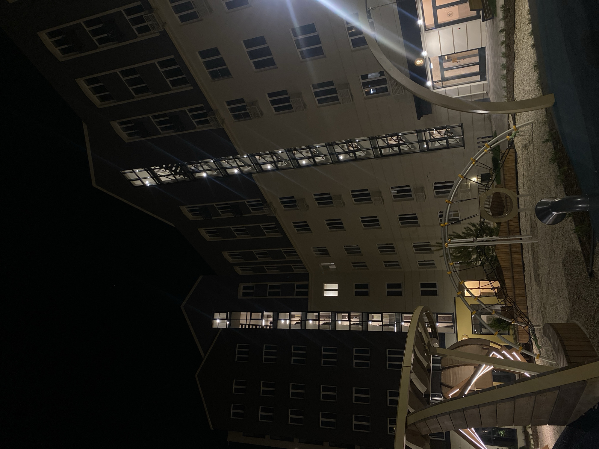 The image depicts a nighttime scene in what appears to be a residential area, likely in Pervomaysky, Russia. A tall building with multiple floors dominates the view, its facade displaying numerous windows, some of which are illuminated, suggesting occupants are home. A connecting bridge-like structure spans between sections of the building. In the foreground, a playground is visible, featuring various play structures, including slides and climbing equipment. The area surrounding the playground is covered with gravel. The lighting is a mix of artificial sources from the building and playground and the ambient darkness of the night. No people are visible in the shot.