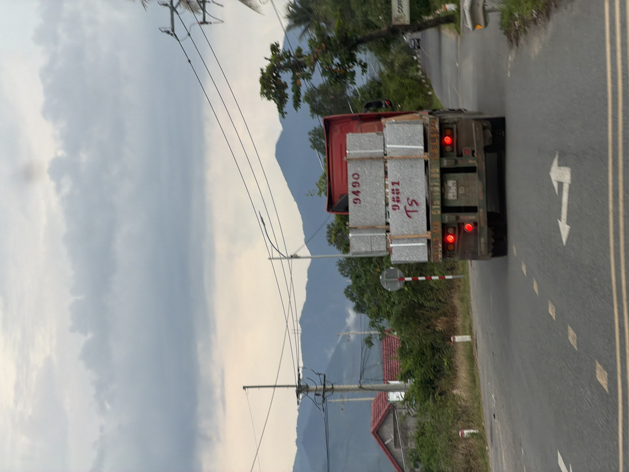 The image shows a red truck with a load of what appears to be granite slabs parked on the side of a road in Diên Thọ, Vietnam. The truck has license plate number "51B.111.95" visible. The granite slabs on the truck are marked with the numbers "9490" and "9881 Ts". The truck is facing away from the viewer and is partially obscured by foliage. In the background, there are mountains, power lines, and houses with red roofs. The sky is overcast with clouds. The road has lane markings and a traffic sign on the right side of the image. There are also some white posts on the side of the road.