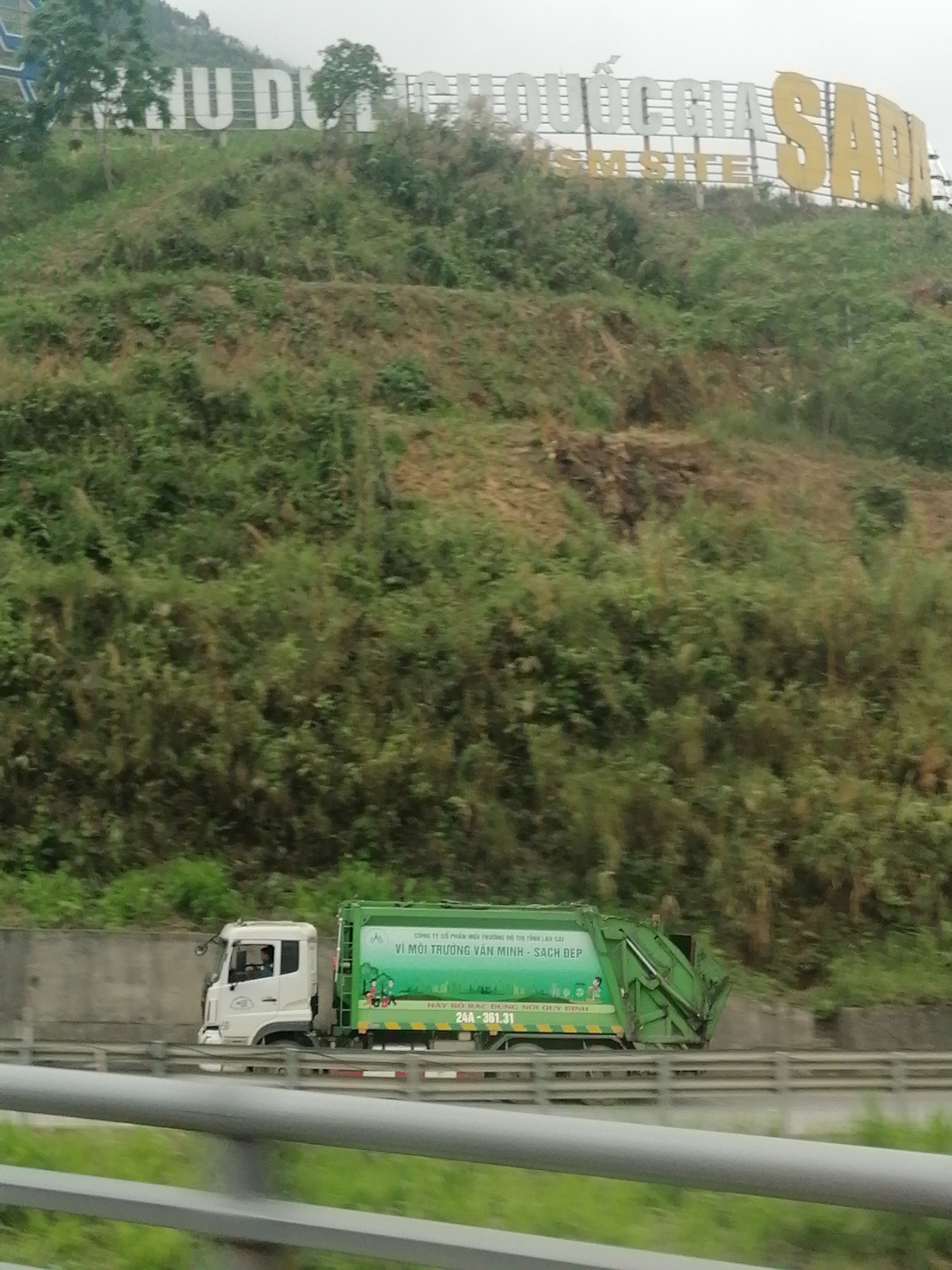 This media file captures a daytime scene featuring a green garbage truck on a road, set against a backdrop of a lush, vegetated hillside. In the foreground, grey guardrails line the side of the road.
The main subject is a white and green garbage truck, seen from the side as it drives on what appears to be a highway or main road. A driver is partially visible inside the truck's cab. The side of the green waste collection unit is covered with Vietnamese text and small illustrations depicting children and nature.
On the hillside in the background, a large sign with prominent white and yellow letters is erected. The white letters partially spell "PHU DU QUỐC GIA," and large yellow letters clearly spell "SAPA." Below "QUỐC GIA," smaller white letters read "SM SITE." The hillside itself is covered in dense green foliage, suggesting a natural or mountainous region.
Visible text on the garbage truck includes:
- "CÔNG TY CỔ PHẦM HỐI THƯƠNG 10 THỊ TỈNH LÀO CAI" (Joint Stock Company for Commerce 10, Lao Cai Province)
- "VÌ MÔI TRƯỜNG VĂN MINH - SẠCH ĐẸP" (For a Civilized - Clean and Beautiful Environment)
- "HÃY BỎ RÁC ĐÚNG NƠI QUY ĐỊNH" (Please put trash in the designated place)
- A numerical sequence, "24A-361.31," likely a license plate or vehicle identification.
The setting appears to be a mountainous or hilly area, likely in Vietnam, given the language and the prominent "SAPA" and "LÀO CAI" references, indicating Sapa, Lao Cai Province. The sky is overcast or hazy, contributing to the soft, diffused lighting. The overall impression is one of a functional vehicle operating within a scenic, natural environment.
