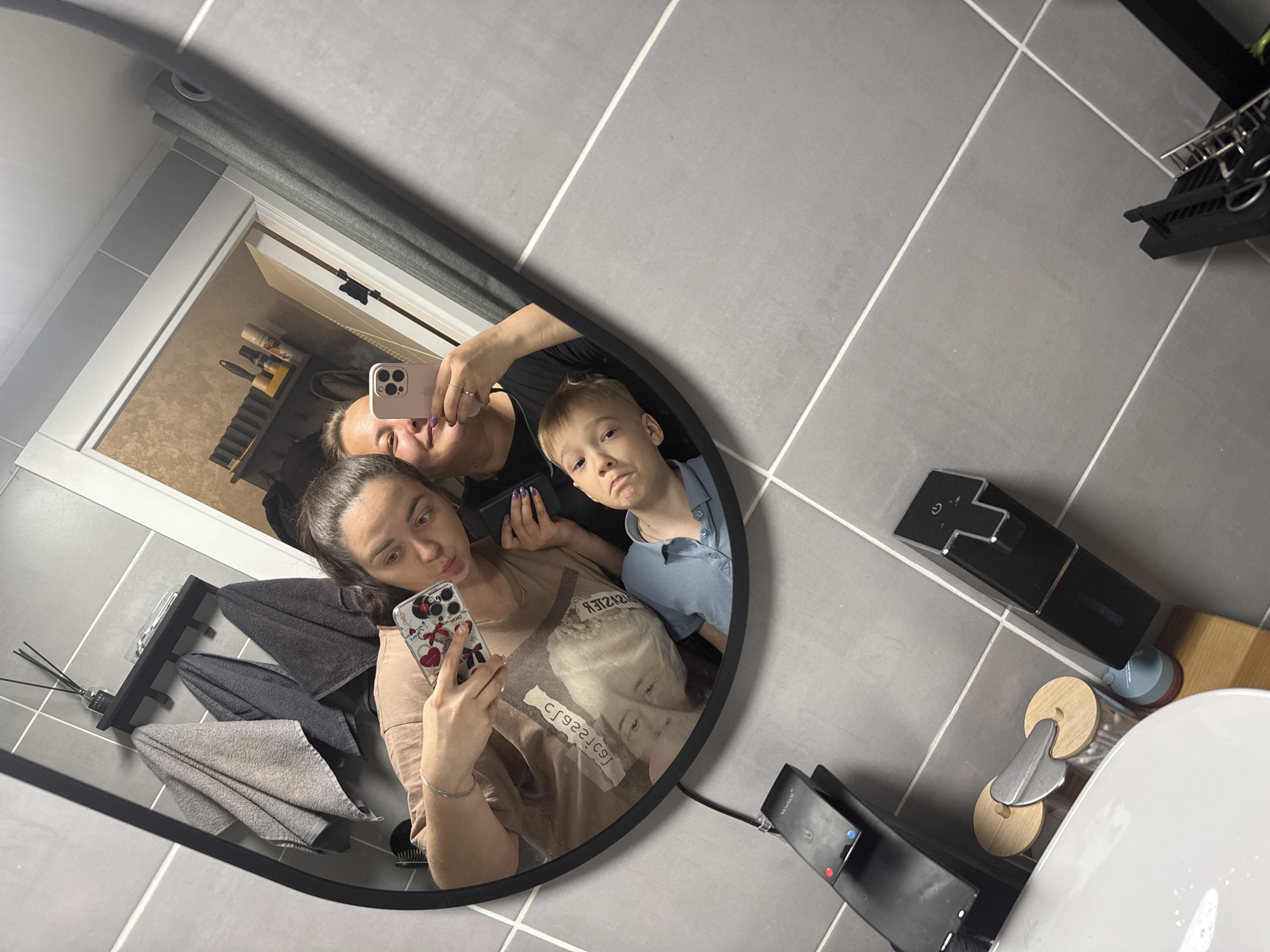 The image shows a reflection in an oval mirror. Three individuals are visible in the mirror: a woman with dark hair, holding a smartphone with a patterned case, appears to be taking a selfie; another woman with blonde hair is seen above her, also holding a smartphone; and a young boy with blonde hair stands to the side.
The setting is a bathroom with gray tiled walls and floor. Various objects are visible in the reflection, including towel racks with towels, toiletries on a shelf, and electronic devices. A toilet and bathroom accessories are also visible. The location cannot be confirmed as Yakhroma, Russia from the provided image.