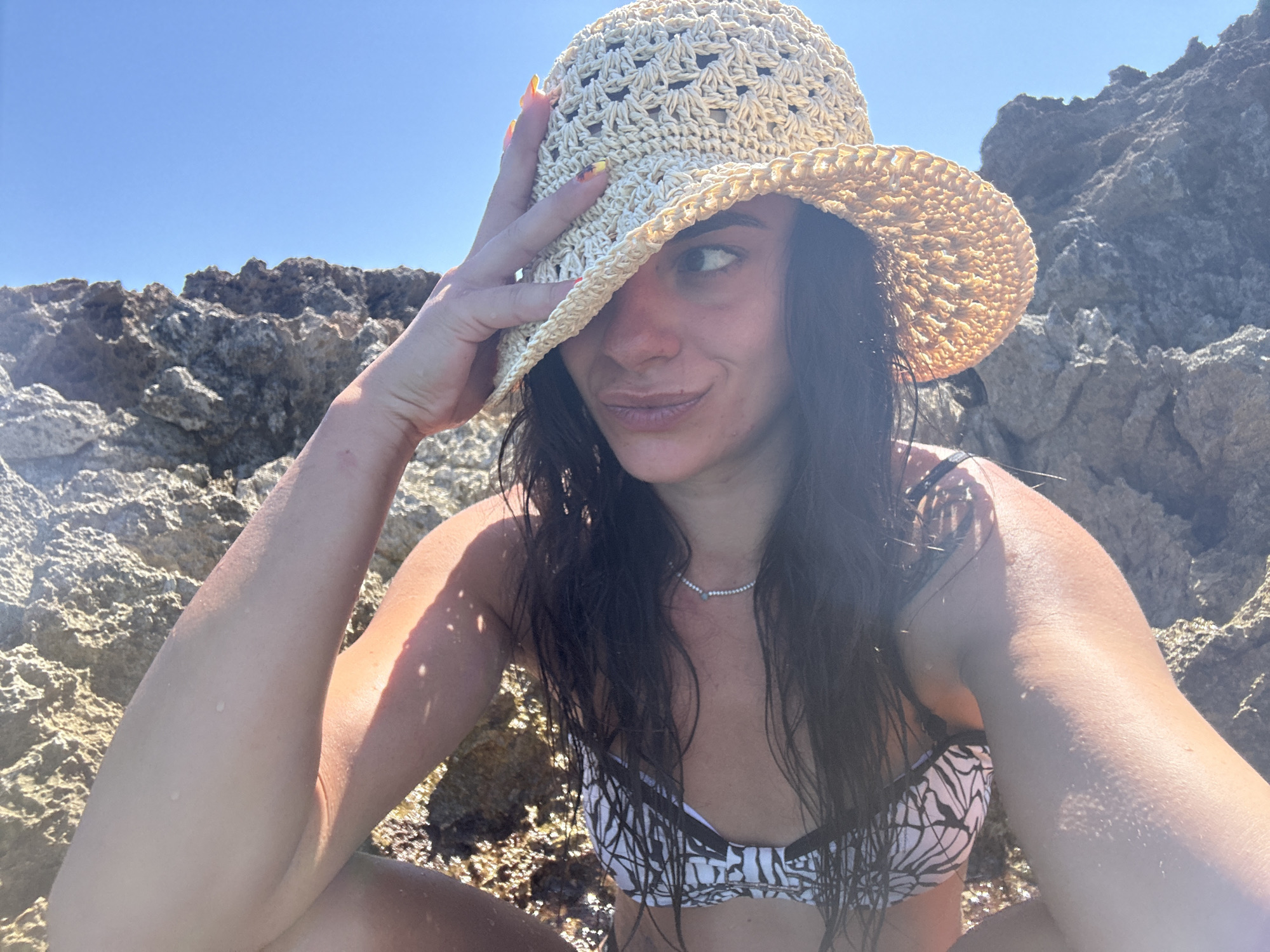 A woman with long, dark, wet hair sits on rocky terrain near the sea, wearing a straw hat and a black and white patterned bikini. She holds her hat with her right hand, obscuring part of her face as she looks towards the viewer. The background consists of sunlit, grey rocks and a clear blue sky. The scene is set in Cala Lliteres, Spain.