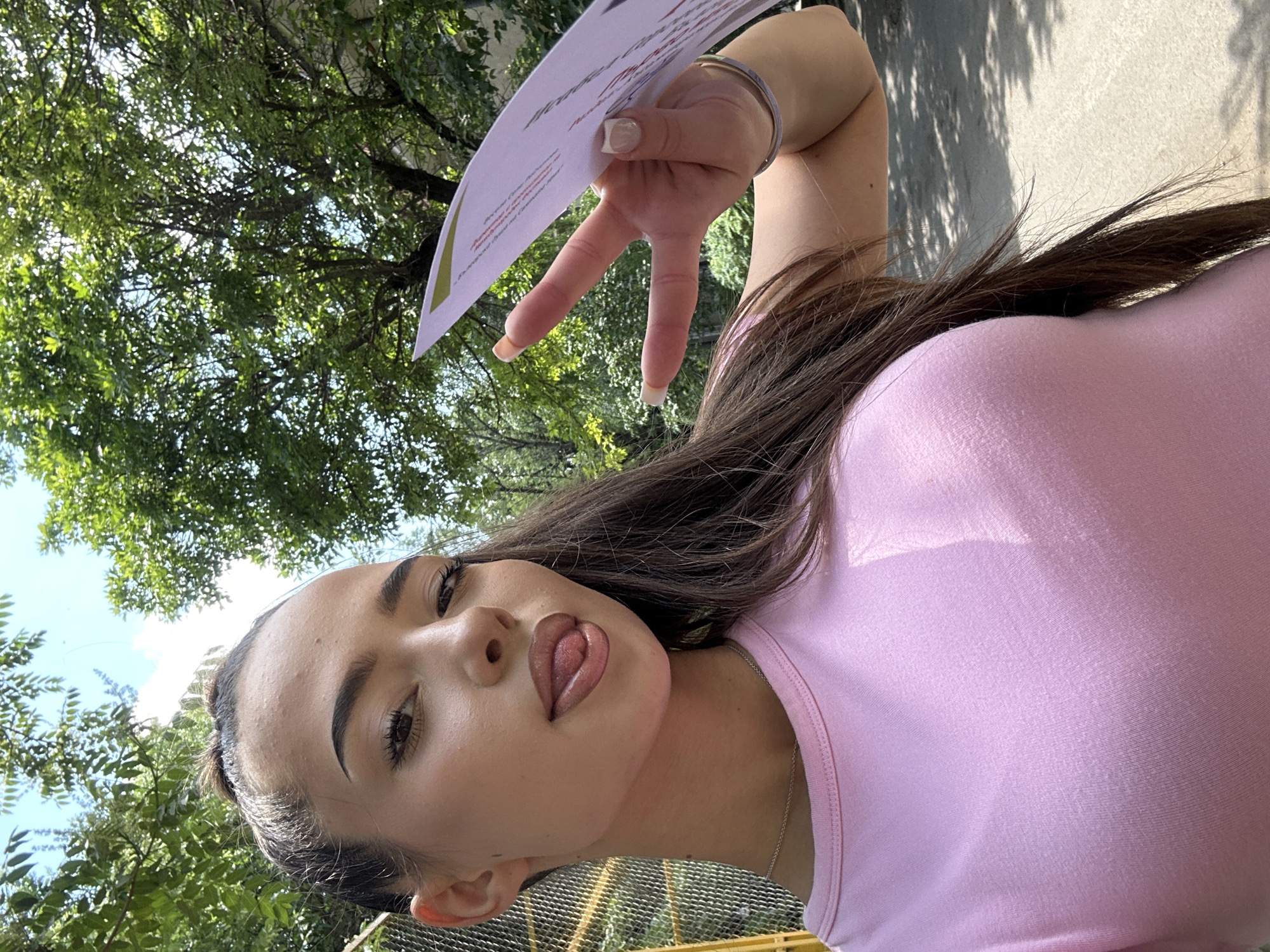 A tilted, low-angle outdoor portrait features a young woman with dark brown hair and a playful expression, sticking her tongue out slightly. She is wearing a light pink top and has visible makeup. Her long, dark hair cascades over her shoulder.
In the upper right of the image, her right hand is raised, making a peace sign (V-sign) with her index and middle fingers. She holds a white piece of paper or flyer, which has some faint, illegible text and possibly a green graphic element. Her nails are manicured with white tips, and she wears a silver-colored bracelet on her wrist and a delicate necklace around her neck.
The background consists of a dense canopy of green leaves and tree branches, suggesting a sunny day in a park or treelined street. Patches of bright blue sky are visible through the foliage. In the lower right background, there appears to be a light-colored fence or barrier. Shadows from the trees are cast onto a wall visible in one of the close-ups. The overall impression is one of a casual, outdoor snapshot on a bright day. No specific text on the paper is legible.