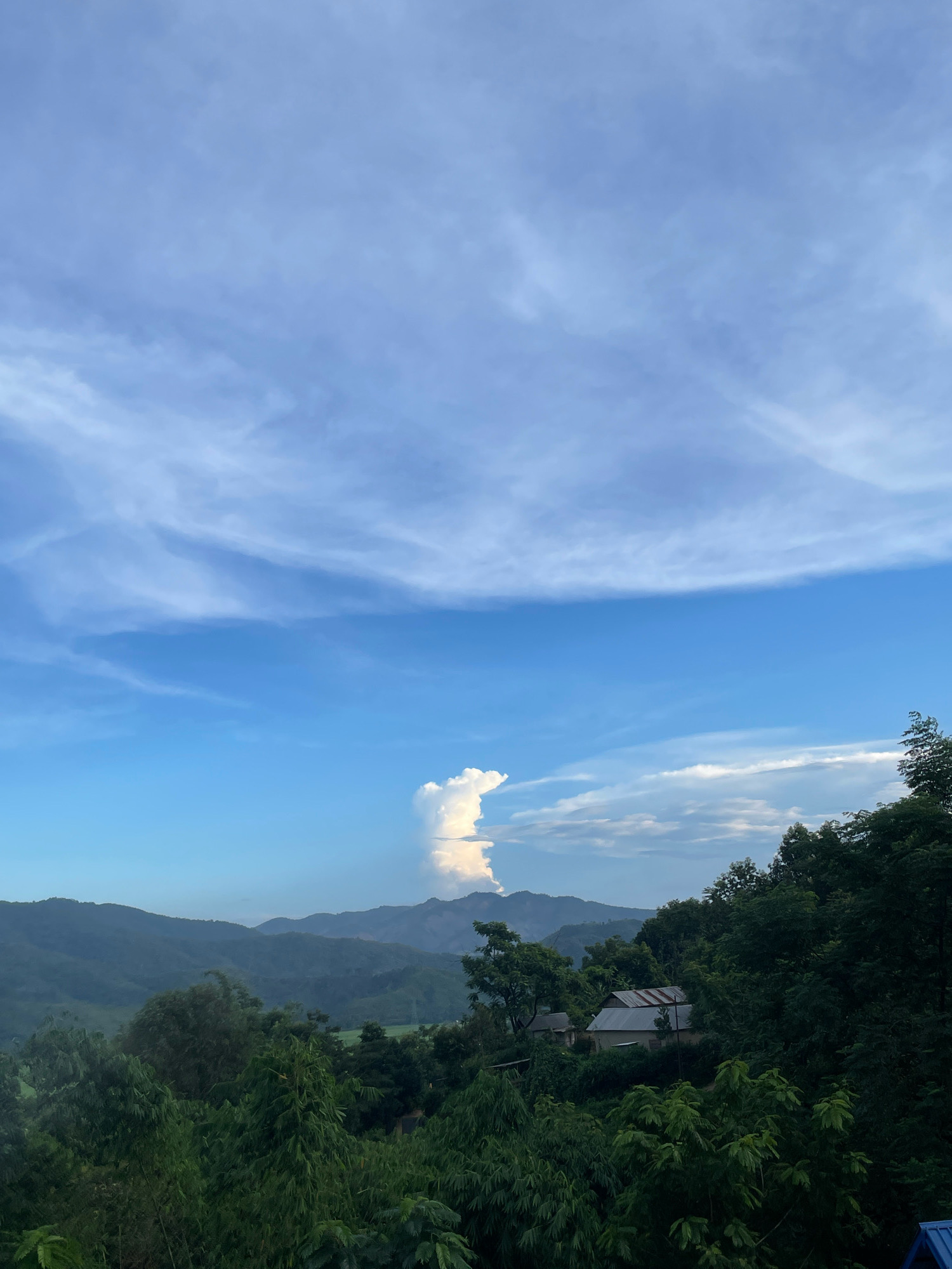 The image depicts a panoramic view of a lush green landscape with rolling hills and mountains under a partly cloudy blue sky. In the foreground, dense foliage of various trees and bamboo covers the hillside, interspersed with a few small houses with corrugated metal roofs. The houses appear to be situated on the slopes, blending into the natural environment. In the mid-ground, layers of mist-covered hills recede into the distance, with a prominent, tall, white cloud formation rising dramatically behind the furthest peaks. The sky is characterized by wispy cirrus clouds streaking across the upper half of the frame, with some cumulus clouds present near the horizon. There are no people visible in the image. The scene suggests a rural or mountainous region, possibly in the Himalayas given the mention of Daili, India.