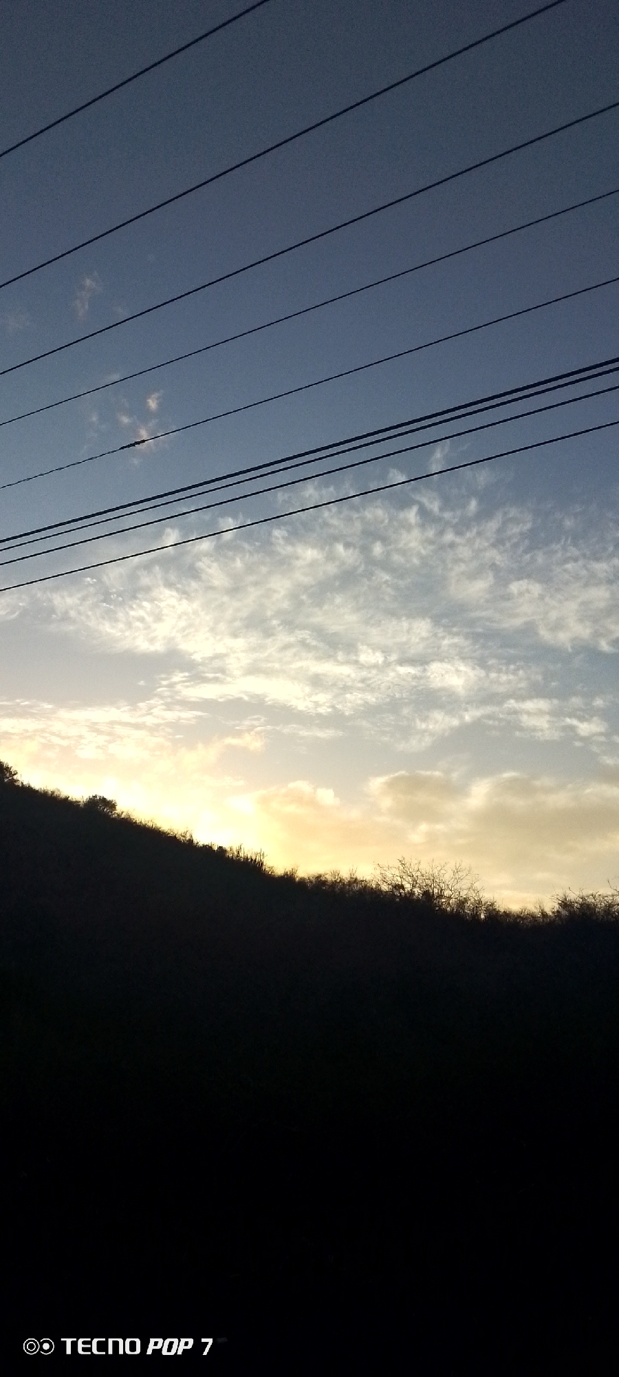 The image captures a landscape at dusk in Altagracia, Venezuela. The lower portion of the frame is dominated by a dark silhouette of a hill or vegetation. Above the silhouette, the sky transitions from a yellow-orange horizon indicating the setting sun to light blue with patches of white clouds. Power lines are stretched across the top of the frame, silhouetted against the sky. The bottom left corner of the image contains the branding for a "TECNO POP 7" mobile phone. No people are visible.