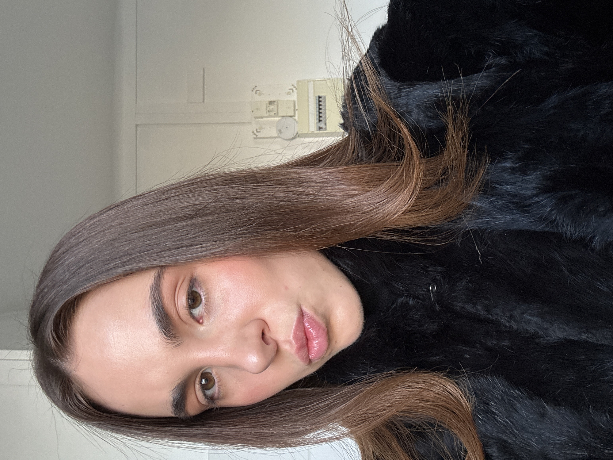 A woman with brown hair is wearing a black fur coat indoors. The location is suggested to be Paris, France, based solely on the user's instructions. The background is a neutral color, featuring a wall with electrical fittings.