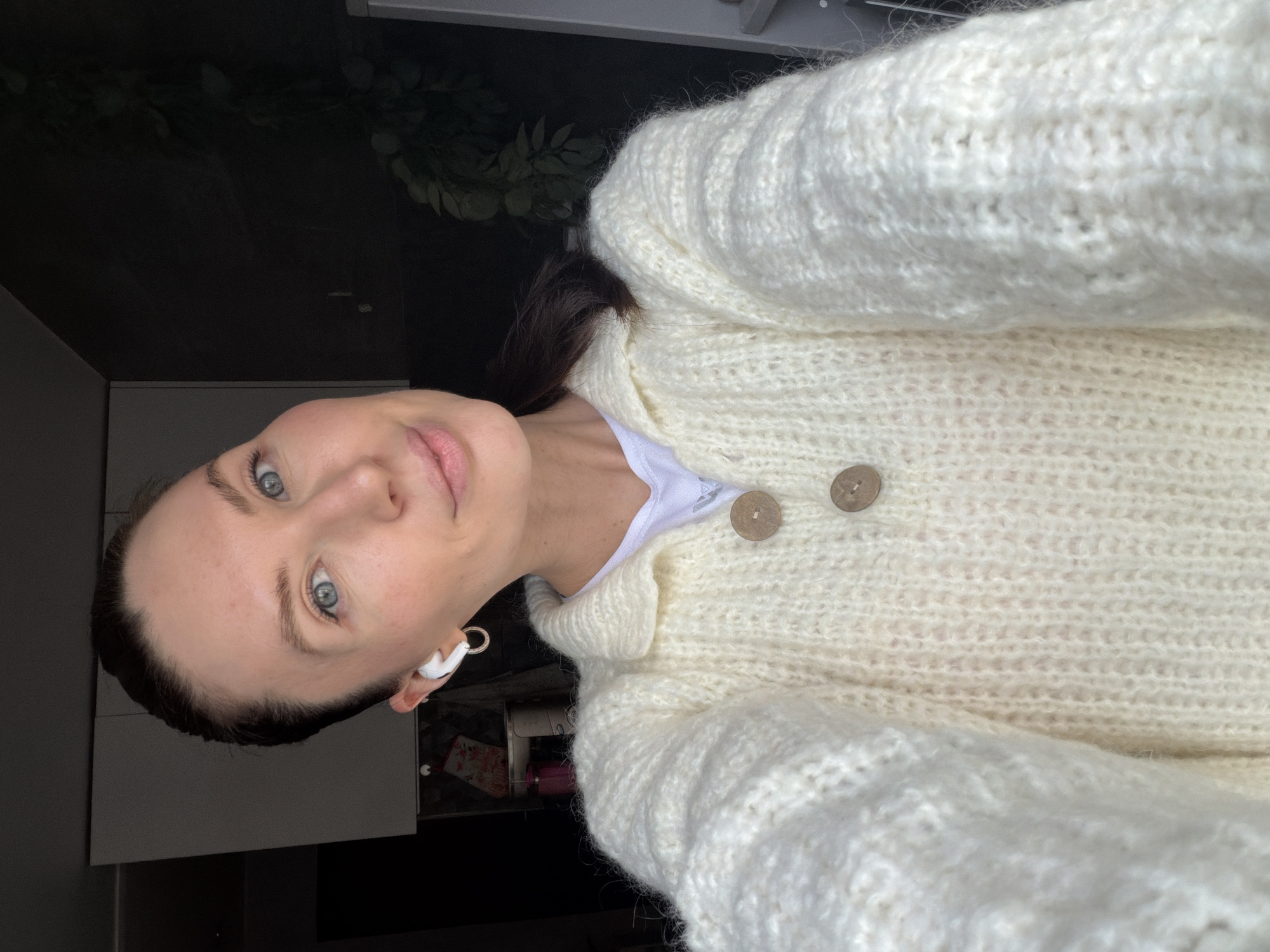 A young adult woman is captured in a close-up, portrait-style selfie, looking directly at the camera with a soft, calm expression and a subtle smile. She has dark hair, fair skin, and striking blue-grey eyes. A white wireless earbud is clearly visible in her right ear.
She is wearing a cozy, cream-colored, chunky knit cardigan or sweater with long sleeves. The sweater is fastened with two prominent, round, light-brown wooden buttons. Underneath, a white t-shirt is visible at the neckline, bearing a partially obscured dark logo or text, possibly a sports brand.
The background is dark and appears to be an indoor setting, likely a home, with hints of modern, dark furniture or cabinetry. Some green foliage, possibly from an indoor plant, is visible in the upper background. In a darker area to the lower left of her face, a floral-patterned phone case and a pink cylindrical object can be faintly seen within a shelf or compartment. The lighting is soft and even, suggesting indoor ambient light rather than bright daylight. The overall impression is one of relaxed comfort.