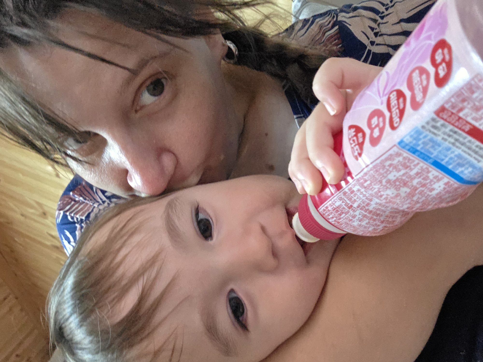 A medium shot captures an overhead perspective of an adult kissing a baby on the cheek. The adult, with brown hair and dark eyes, is wearing a blue and white patterned shirt and has an earring in their left ear. The baby, with brown hair and wide dark eyes, is facing the camera and appears to be drinking from a pink bottle with Asian text and heart shapes printed on it. The background consists of wooden paneling. The scene takes place in a residential setting in Dunnae-myeon, South Korea.