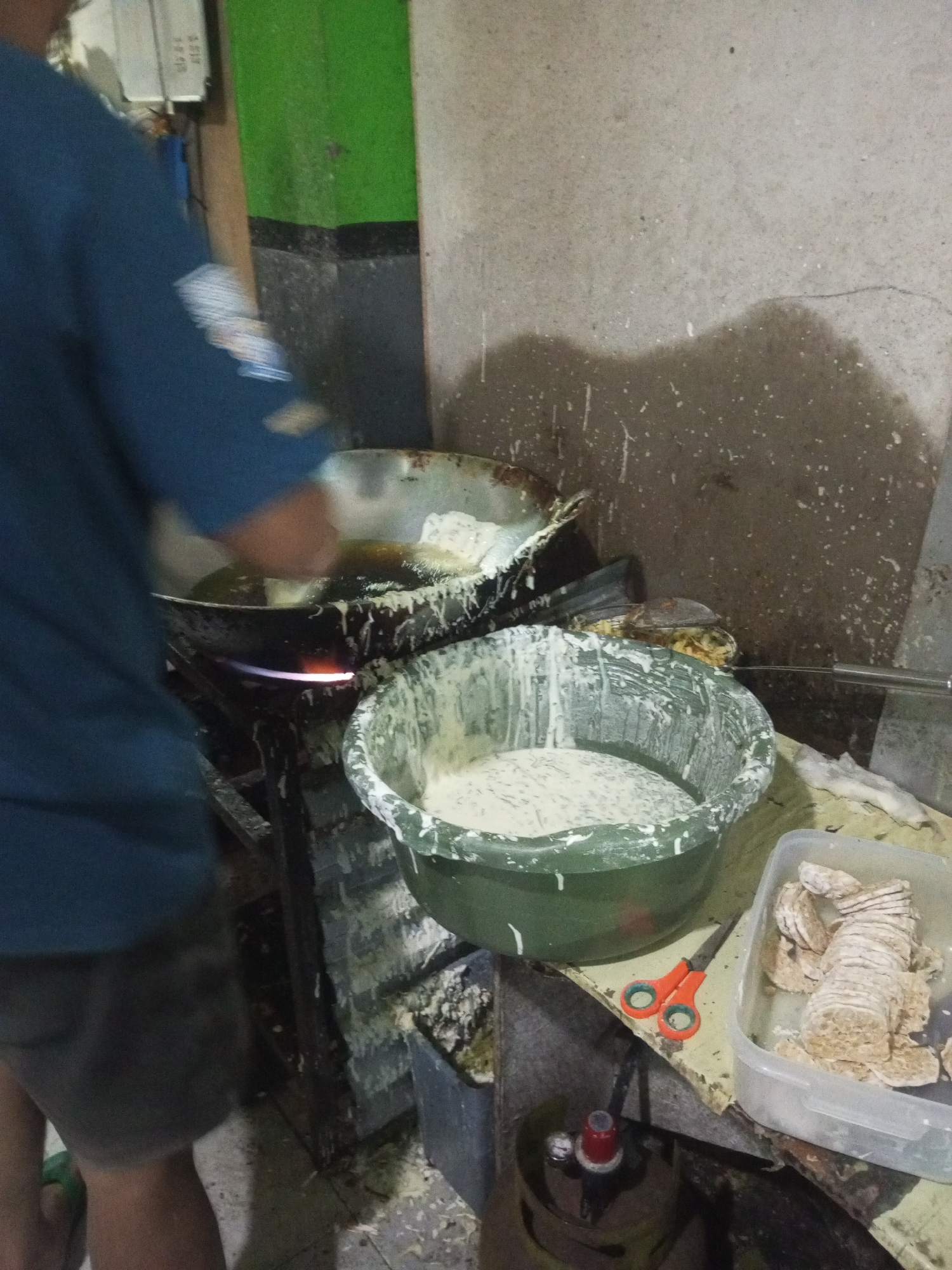 The image depicts a cooking scene, likely outdoors or in a semi-enclosed space, with a focus on food preparation. In the foreground, a large green bowl is filled with a white batter or mixture. To the right of the bowl, a clear plastic container holds several pieces of what appears to be tempeh, a fermented soybean cake commonly consumed in Indonesia. A pair of orange-handled scissors rests on the table next to the bowl.
Dominating the mid-ground is a large, dark wok or pan being heated by a visible flame from a gas burner. A person, mostly obscured by a blue shirt and dark shorts, is engaged in cooking in the wok. Their hands, covered in batter, are actively involved in frying food items submerged in hot oil. The wok is splattered with oil and batter, indicating ongoing cooking.
In the background, a textured wall with dark stains and white splatters suggests a working environment, possibly a kitchen or food stall. A green-painted vertical surface with a black stripe is also visible. An electrical outlet is mounted on a white panel in the upper left corner. The overall impression is of a busy food preparation area, likely a small business or household operation in Andongsili, Indonesia, given the contextual information. The lighting is dim, and the image has a slightly grainy quality, suggesting it was taken in low light conditions.