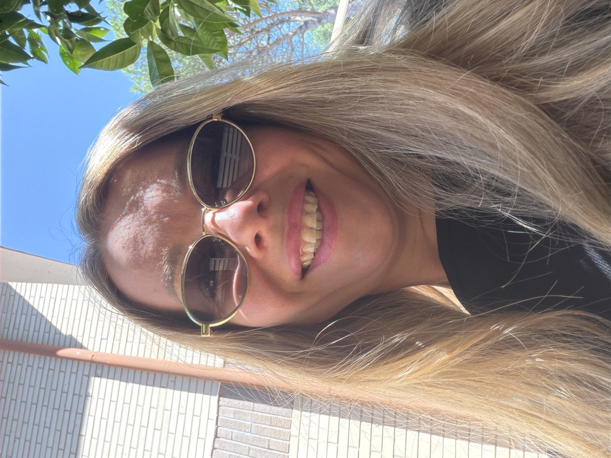 The image is a close-up selfie of a woman with blonde hair and sunglasses, taken outdoors on a sunny day. She is wearing a black top. In the background, there is a brick wall with a downspout, trees, and a clear blue sky. The lighting suggests the sun is overhead, casting shadows. The overall setting appears to be casual and outdoor, possibly a residential area or a park. There is no visible indication of the city of Rome, Italy, or any other specific location.
