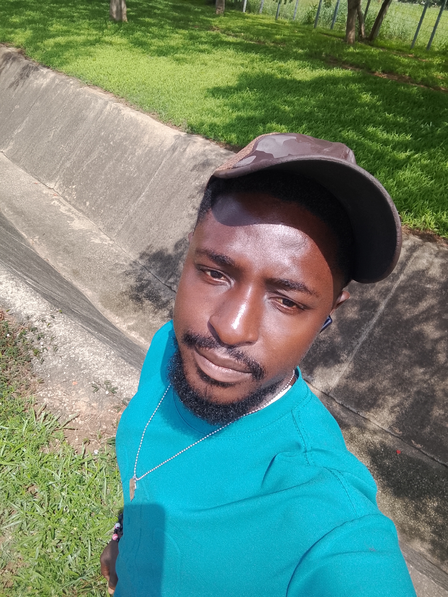 An adult male, approximately 20-30 years of age, with a dark complexion and a trimmed beard, is depicted in an outdoor setting in Karmajiji, Nigeria. He wears a dark, possibly camouflage-patterned baseball cap, a teal-colored long-sleeved shirt, a thin silver chain with an elongated brown or wooden pendant, and a dark earbud in his right ear. Bright sunlight illuminates his face, creating strong highlights and shadows. The subject is looking directly into the camera, positioned from a slightly elevated angle, suggesting a selfie. The immediate background features a weathered concrete drainage channel or retaining wall. Beyond the concrete, vibrant green grass is visible, interspersed with patches of shade. The distant background reveals trees and a wire mesh fence. The overall environment is sunny and semi-natural, characteristic of an open area or park.