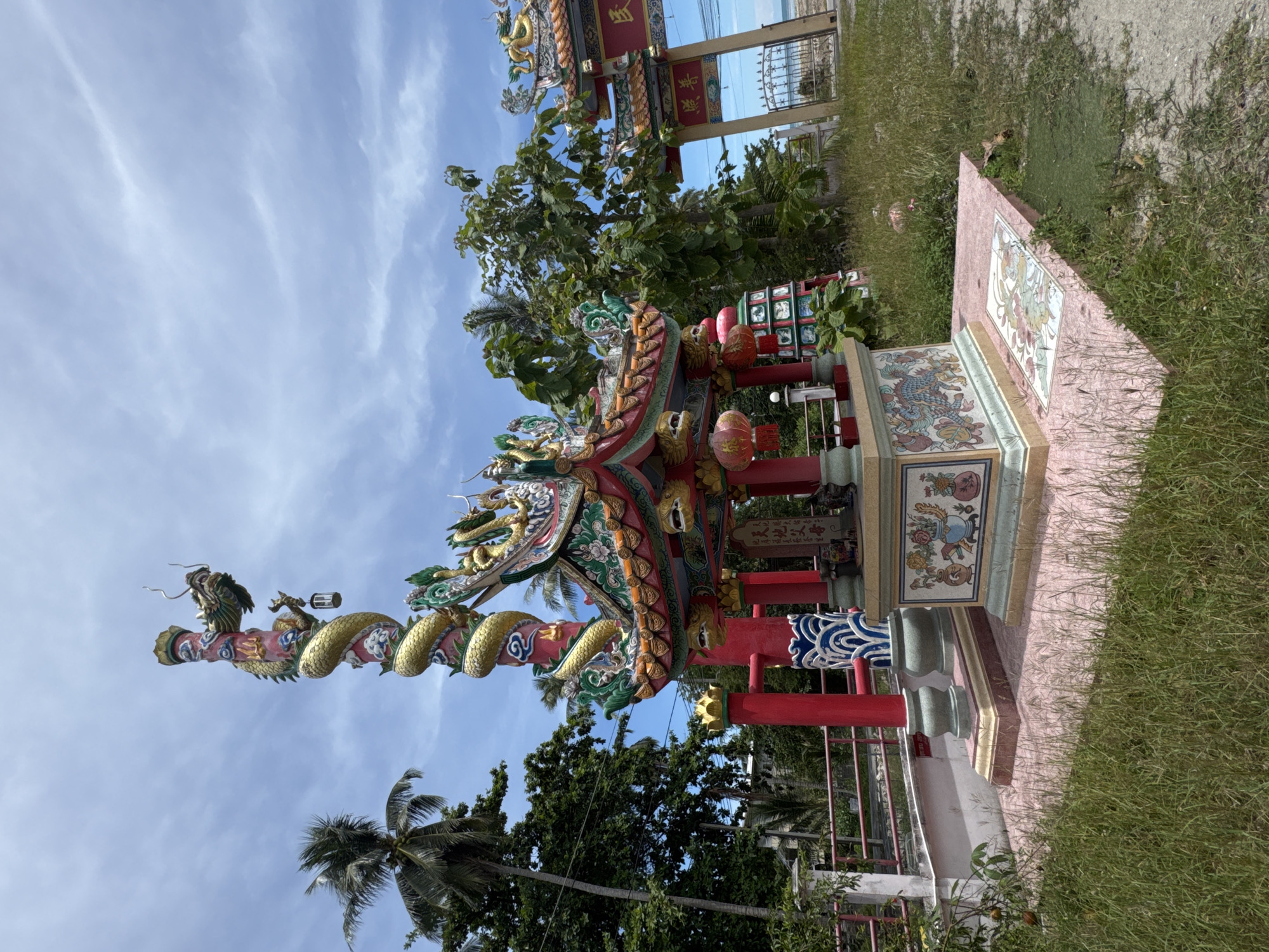 The image shows a small, ornate Chinese temple or shrine in Koh Phangan, Thailand, on a clear day with a partially cloudy blue sky. The structure is vibrant with red, gold, green, and blue accents. A prominent feature is a tall, red pillar wrapped by a golden dragon sculpture. In front of the pillar is the main shrine building, characterized by a multi-tiered, colorful roof adorned with dragon figures and decorative eaves. Below the roof, there are several smaller, red lanterns hanging. The base of the shrine is decorated with colorful tile panels depicting dragons and floral motifs. The shrine is situated on a pink tiled platform, surrounded by overgrown grass and some vegetation. To the right, a large arched gateway, also decorated in a Chinese style with red and gold, stands open, revealing a glimpse of the ocean and a fence. The setting appears to be outdoors, possibly near a coastal area, given the visible ocean in the background. There are no people present in the image.