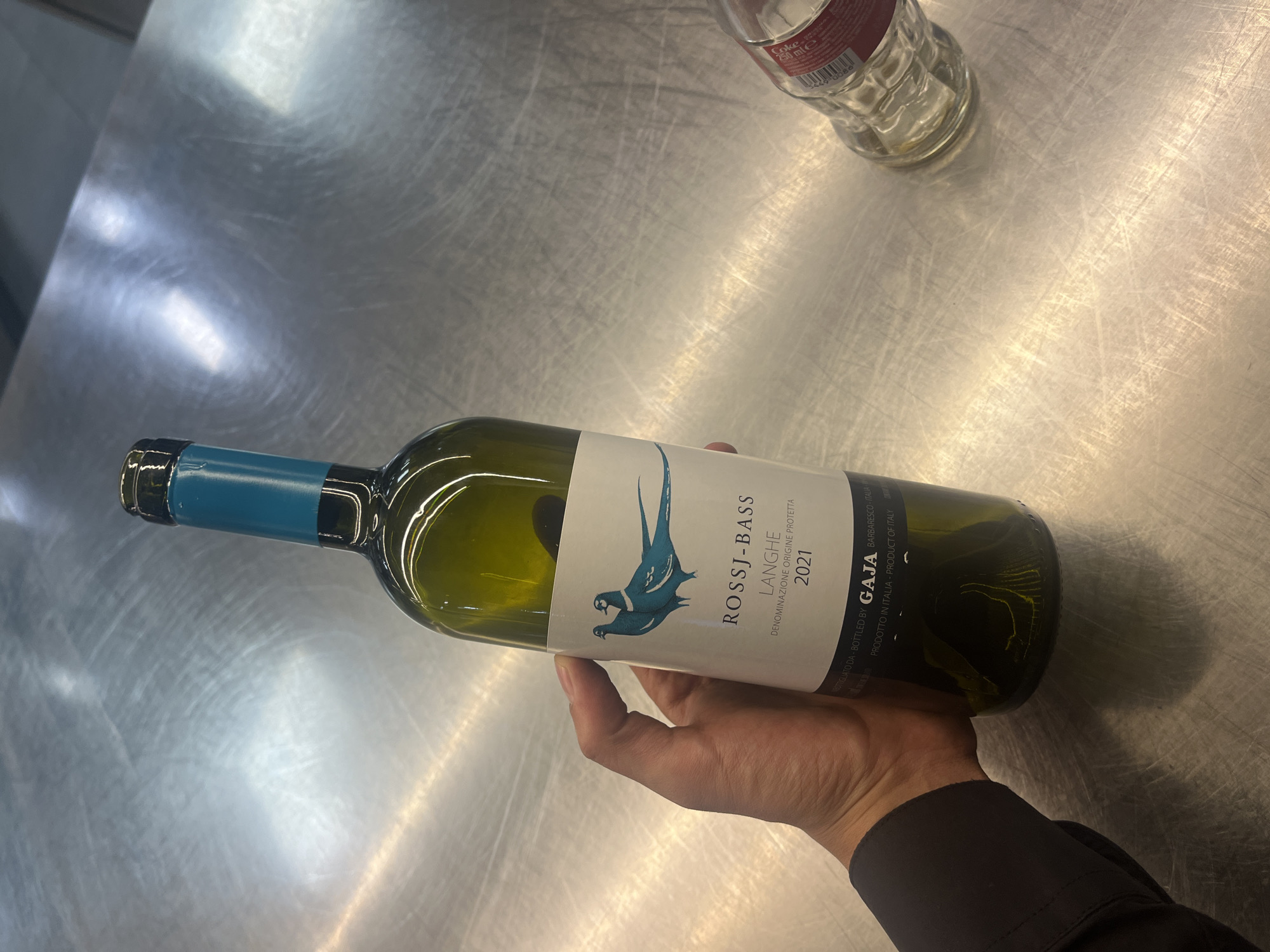 A person's hand holds a wine bottle horizontally over a reflective, scratched metallic surface. The wine bottle is made of green glass with a prominent white label. The label features an illustration of two blue pheasants and clearly displays the text "ROSSJ-BASS," "LANGHE," "DENOMINAZIONE ORIGINE PROTETTA," and the vintage "2021." Below this, smaller text reads "BOTTIGLIATO DA - BOTTLED BY GAJA BARBARESCO - ITALIA -" and "PRODOTTO IN ITALIA - PRODUCT OF ITALY." The bottle's neck is covered by a blue foil or cap.
In the upper right corner of the image, partially visible, is a clear plastic bottle, identified by the text "Coke" and "250 ml" on its label. The metallic surface, which appears to be a countertop or table, shows numerous scratches and reflections, indicating an indoor setting with overhead lighting. The exact time of day and weather are not discernible. There are no specific visual cues in the image to confirm the location as Sofia, Bulgaria. The activity depicted is simply the holding or presentation of the wine bottle.