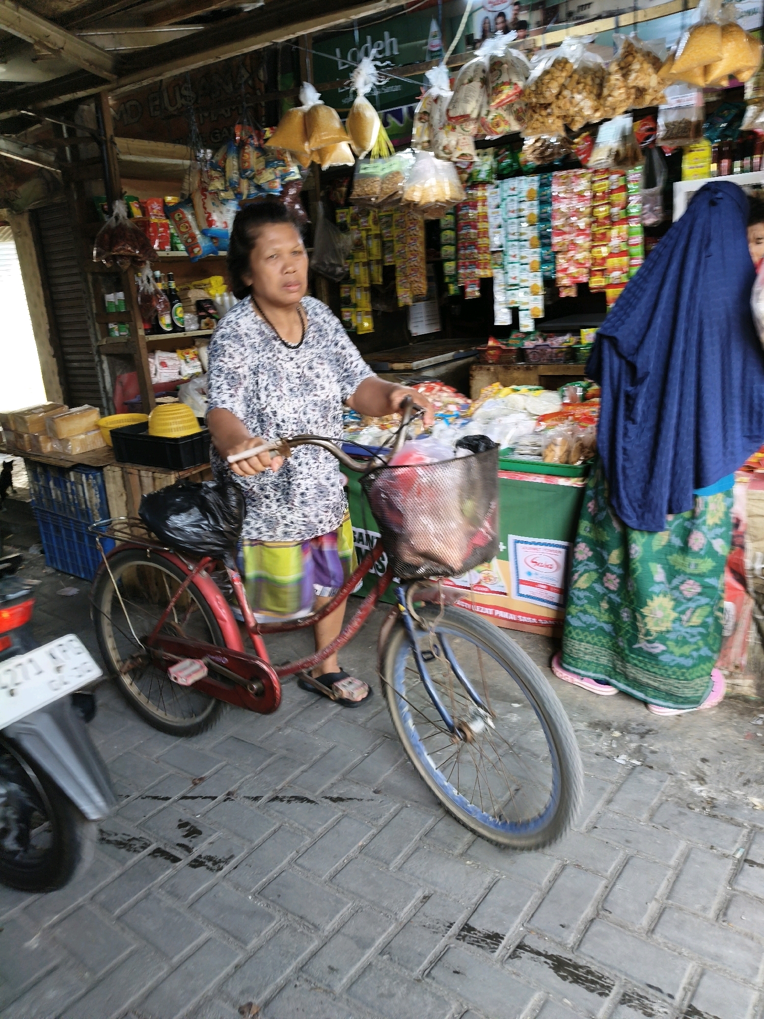 In Setiamekar, Indonesia, a woman stands next to a bicycle at a street-side market. She is wearing a patterned blue and white shirt and a colorful striped skirt. Her right hand is on the handlebars of the bicycle, which has a wire basket attached to the front. A black plastic bag is secured to the rear rack of the bicycle. Behind the woman, shelves are stocked with various packaged goods, including snacks and drinks. A second person, largely obscured by a dark blue fabric, is visible to the right of the woman. The market appears to be outdoors, with a wooden structure overhead and a paved ground surface.