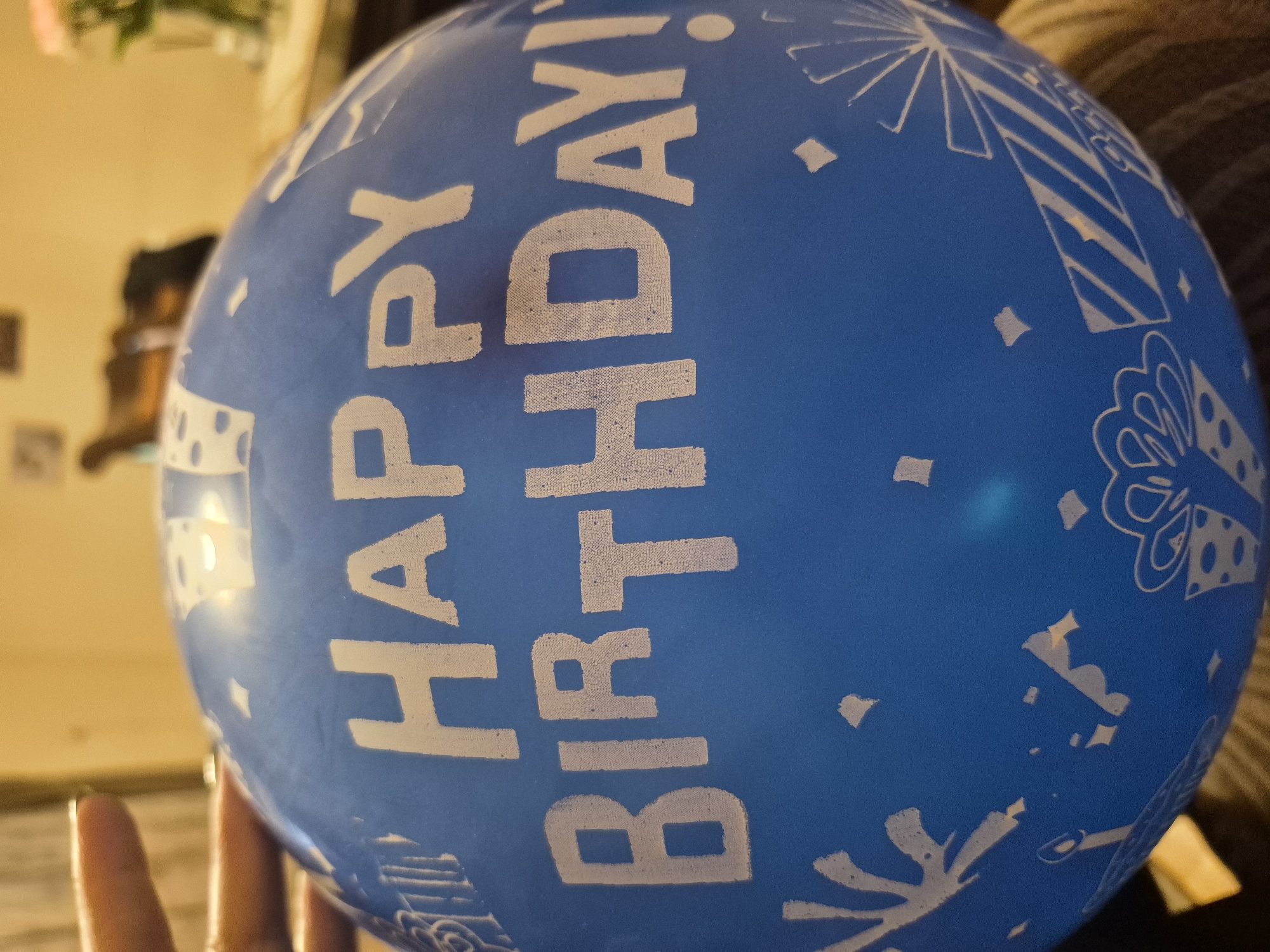 The image shows a blue birthday balloon held in a person's hand indoors. The balloon has white text that reads "HAPPY BIRTHDAY!" vertically aligned. The balloon also features various white birthday-themed illustrations, including gift boxes and confetti. The background is blurred and includes glimpses of a room interior with furniture and a light-colored wall. This image doesn't contain information that leads to identifying the location.