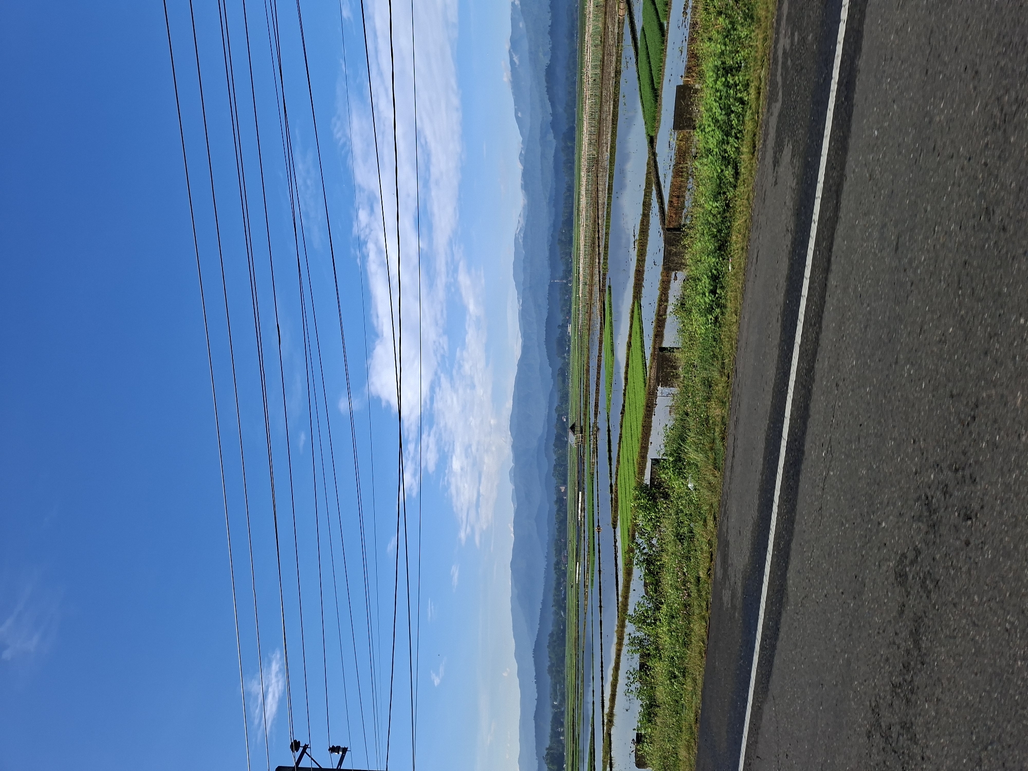 The image depicts a rural landscape in Kampung Jumpa, Indonesia. A paved road with a white line runs along the bottom of the frame. To the right of the road, lush green vegetation borders flooded rice paddies. The paddies are geometrically arranged and reflect the bright blue sky, which is dotted with wispy white clouds. In the distance, a range of hazy blue mountains forms the horizon. A small, traditional hut with a thatched roof is visible within the rice fields. Overhead, several power lines are strung across the sky, supported by a utility pole on the left side of the image. The scene is bathed in natural daylight, suggesting an outdoor setting. There are no people or animals visible in the image.