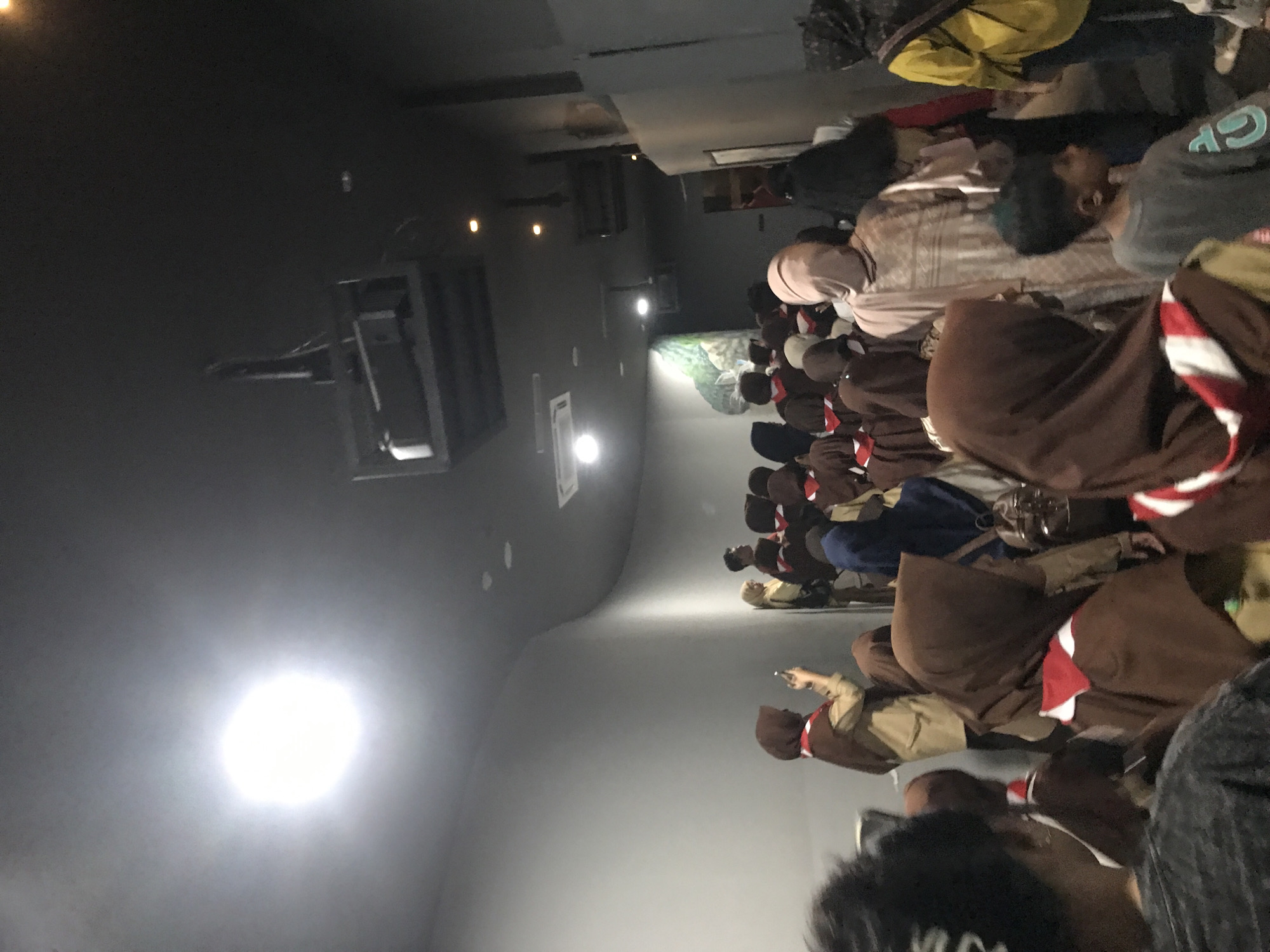 A group of people, mostly wearing brown and white head coverings and brown clothing with red and white neckerchiefs, are seated in rows in a dimly lit room. There is a projector and lighting fixture on the ceiling, and a light source is on the left. This scene possibly takes place in Banda Aceh, Indonesia, as suggested by the user. The nature of the gathering and the activities taking place are not explicitly evident in the image. One person appears to be using a mobile device.