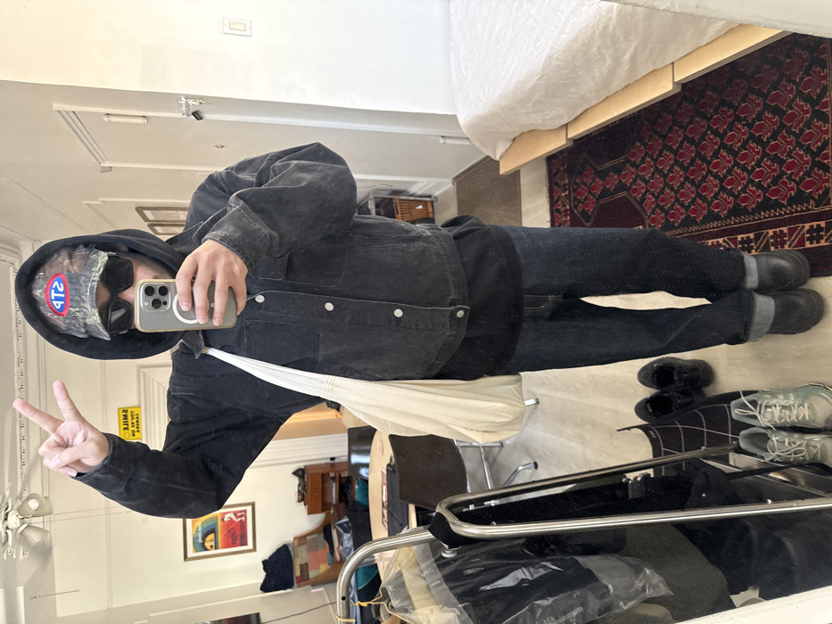 A full-body mirror selfie captures an adult individual standing within a residential interior in New York, United States of America. The subject is dressed in a dark, possibly charcoal-colored, hooded denim jacket with light buttons, a black t-shirt, dark blue jeans cuffed at the ankles, and black shoes. They wear a camouflage baseball cap with a red oval "STP" logo, and dark sunglasses. A white fabric tote bag is slung over their left shoulder. The individual holds a light-colored smartphone with a visible circular magnetic attachment in their right hand, using it to capture the reflection. Their left hand is raised, forming a "peace" sign.
The immediate environment visible in the reflection includes a light-colored wooden floor partially covered by a dark red patterned rug with geometric motifs. To the left of the subject, a metal clothing rack with garments draped in plastic is visible, alongside a round wooden table with chairs, a dark wooden cabinet, and a framed artwork on the wall. A yellow rectangular sign bearing the mirrored text "SMILE YOU ARE ON CAMERA" is also present on the left wall. Two additional pairs of shoes, grey sneakers and black slip-ons, are visible on the floor near the rack. To the right, a minimalist light-colored wooden bed frame with a white mattress and white bedding is positioned. A white door with a silver handle and a light switch are visible on the far right wall. A ceiling fan with light fixtures is reflected in the upper left portion of the mirror.