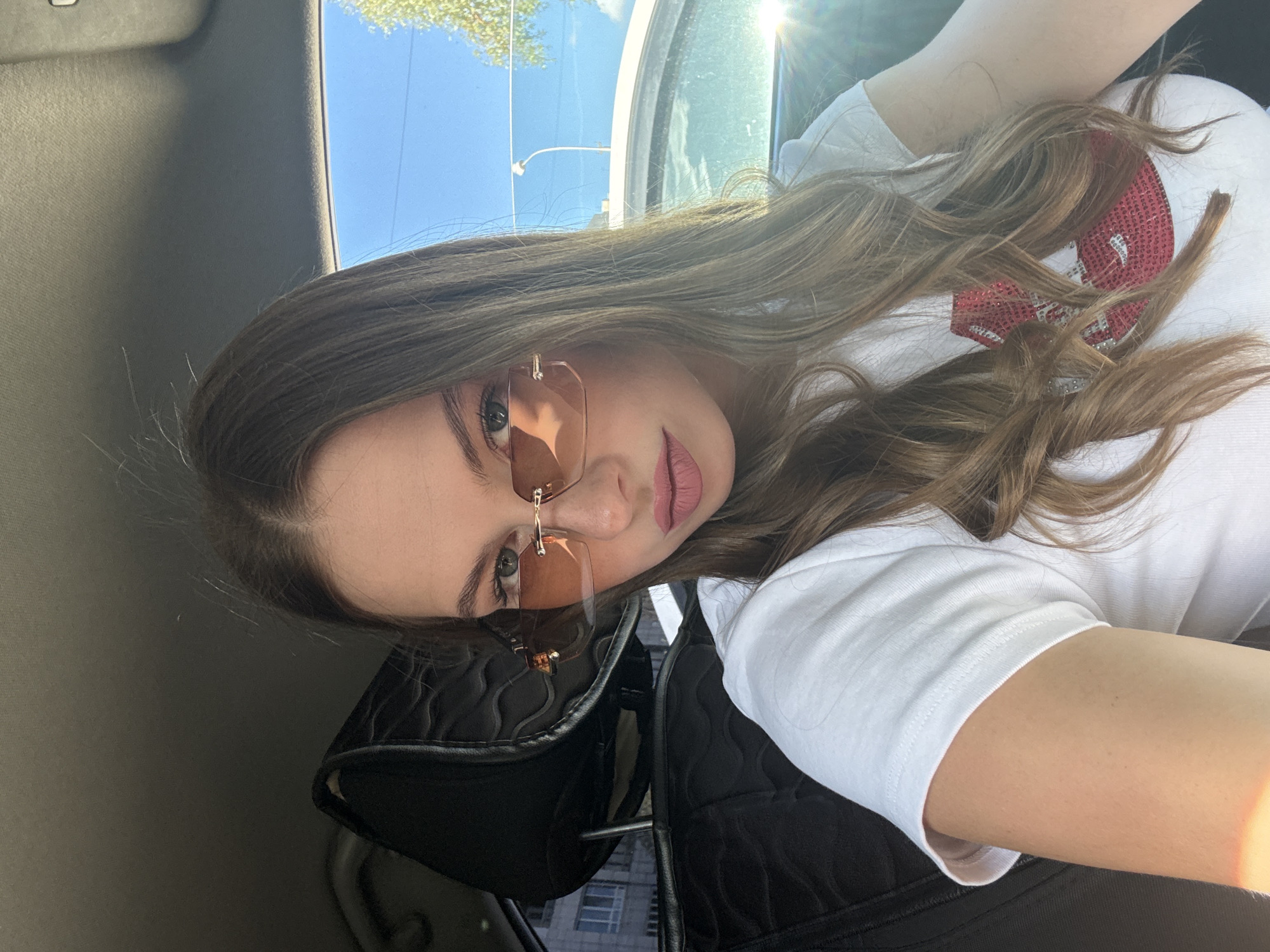 A young woman with long, wavy brown hair and fair skin is seated in the driver's seat of a car. She is wearing sunglasses with pinkish-orange lenses and a white t-shirt with a red sequined design on the chest. Her lips are painted a pale pink. The interior of the car, with a black headrest and dark upholstery, is visible. Outside the car window, a clear blue sky and trees can be seen, along with a streetlamp and a building in the background. The sun is shining brightly, casting a glare on the car's interior. The photograph was taken in Perm, Russia.