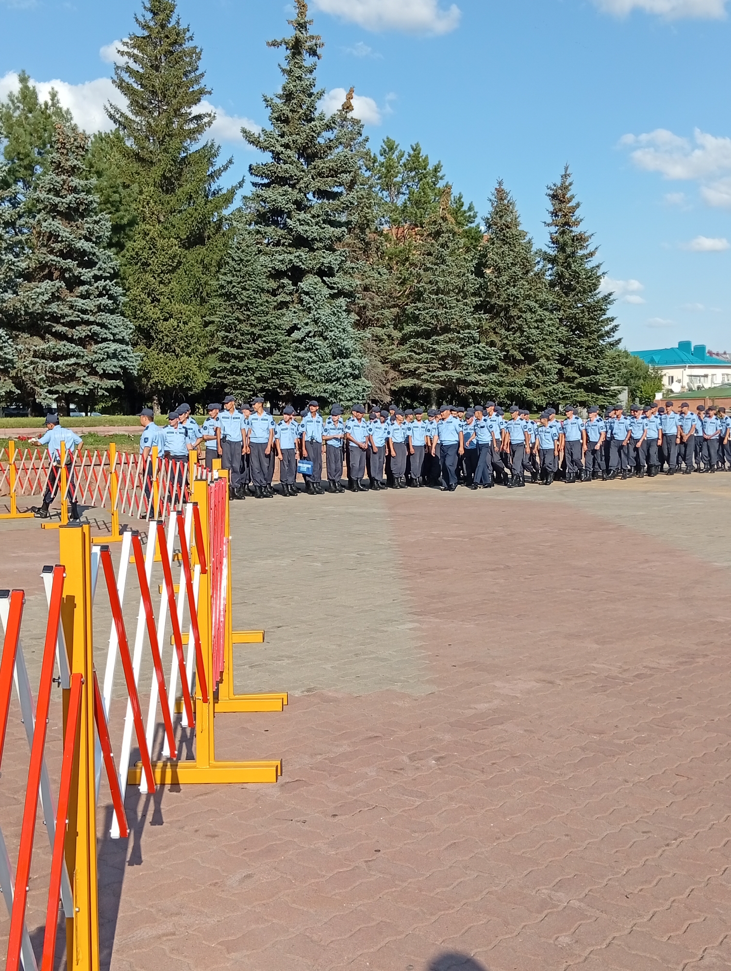 A long line of people in light blue uniforms and dark caps stand at attention outdoors on a paved area, with a row of red, white, and yellow retractable barriers in the foreground. The individuals appear to be male and are in formation. Behind them, a line of tall evergreen trees forms a backdrop against a bright blue sky with scattered white clouds. To the far right, a light blue-roofed building with white walls is visible in the distance. This scene is on the territory of the Kostanay Academy of the Ministry of Internal Affairs of the Republic of Kazakhstan, located in the city of Kostanay, Kazakhstan.