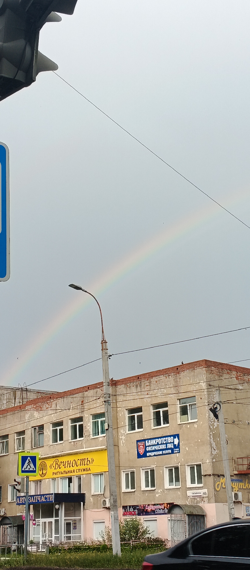 A faint rainbow arcs across a grey, overcast sky above a multi-story building in Izhevsk, Russia. The building appears aged, with a weathered, light-colored facade and a brick parapet. Several windows with white frames are visible. Signs are affixed to the building's facade; one yellow sign reads "Вечность" (Eternity) with "РИТУАЛЬНАЯ СЛУЖБА" (Ritual Services) below it, featuring a tree graphic. Another blue sign displays "БАНКРОТСТВО ФИЗИЧЕСКИХ ЛИЦ ЮРИДИЧЕСКИЕ УСЛУГИ" (Bankruptcy for Individuals Legal Services). A street light on a pole stands in front of the building. In the foreground, a black car is partially visible. A pedestrian crossing sign is also present. The overall scene suggests a typical urban environment under cloudy weather conditions.