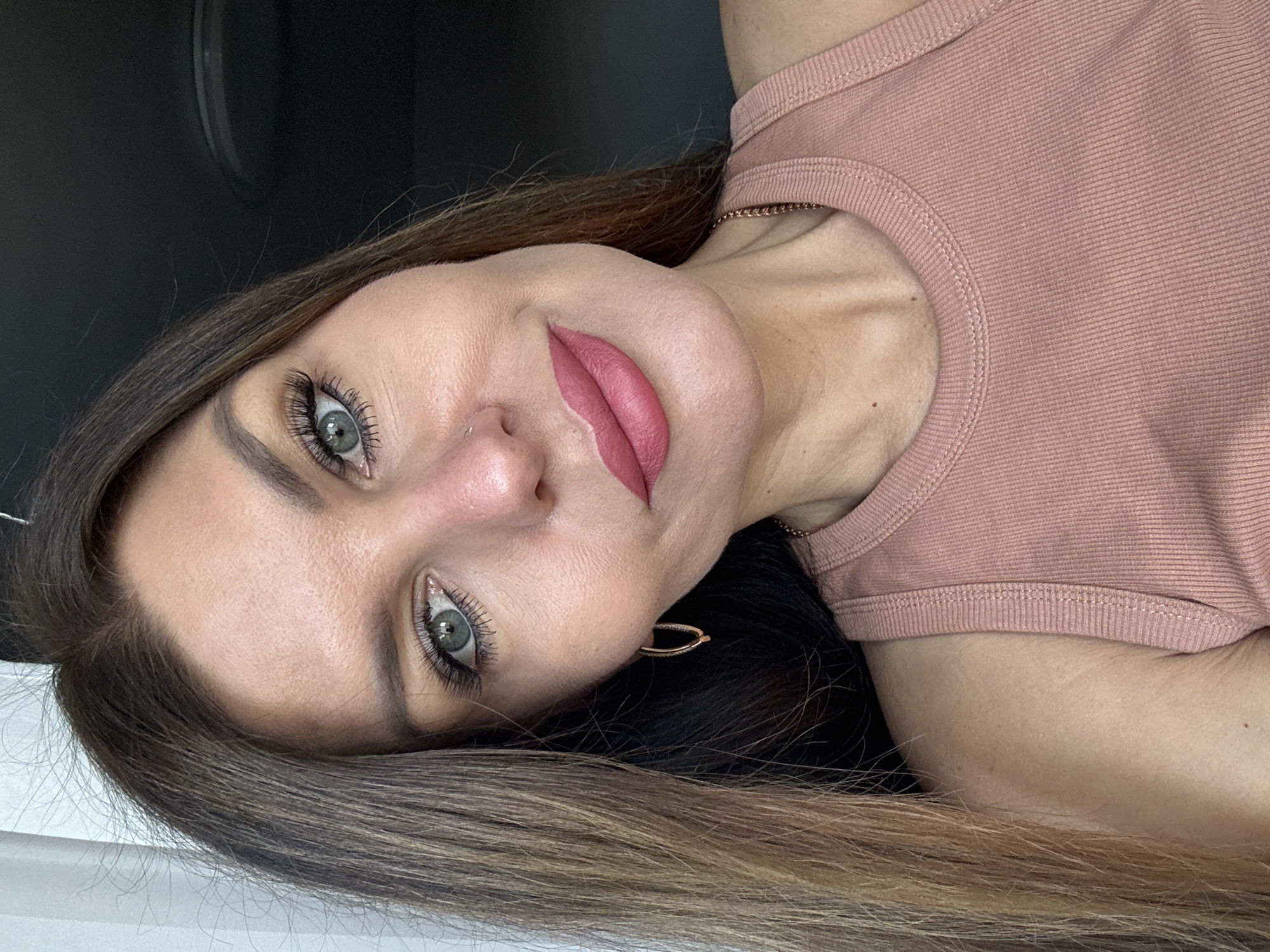 A close-up selfie of a woman with long brown hair, green eyes, and full lips. She is wearing a pink tank top and gold hoop earrings. Her makeup includes eyeliner, mascara, and pink lipstick. The background is dark. The image was taken in the city of Chaikovsky, Russia.