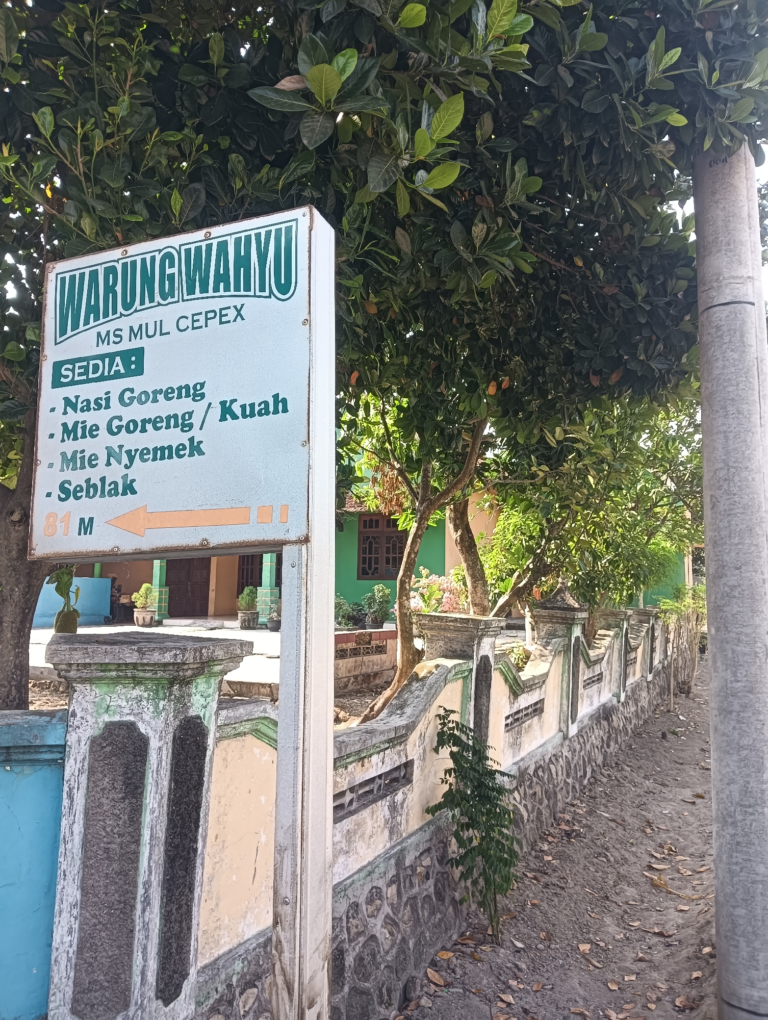 The image captures a street-level view of a sign for "WARUNG WAHYU MS MUL CEPEX" located in Tanggan, Indonesia. The sign, prominently displayed on the left, is rectangular with a green border and features white text. The top section reads "WARUNG WAHYU" in large, stylized green lettering. Below this, "MS MUL CEPEX" is written in smaller, uppercase green text. The sign then lists menu items in green bullet points: "Nasi Goreng," "Mie Goreng / Kuah," "Mie Nyemok," and "Seblak." At the bottom left of the sign, an orange arrow points to the left, indicating a distance of "81 M."
The sign is partially obscured by the dense green foliage of a large tree with dark green leaves, likely a jackfruit tree, with some light orange fruits visible. In the background, a low, stone and plaster wall runs along the right side of the frame, painted in alternating light green and cream colors, with stone detailing at the base. Behind the wall, a building with green walls and a brown roof is visible, featuring traditional Indonesian architecture with wooden window frames.
The ground in front of the wall is a mix of dirt and fallen leaves, with a small, leafy plant growing near the wall. A light gray concrete utility pole stands to the right of the sign, partially in the frame. The overall scene suggests a modest, local establishment in a tropical environment. The image appears to have been taken during the day, with natural light illuminating the scene.
