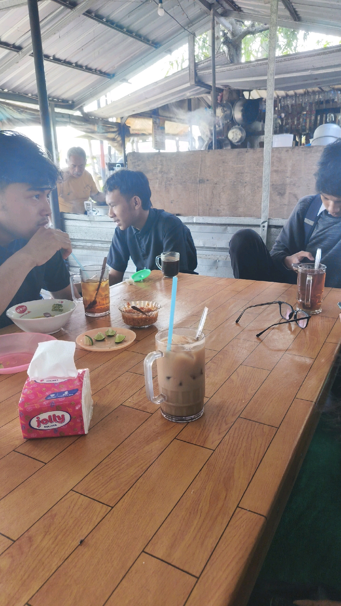 The image depicts an outdoor dining scene at what appears to be a food stall or small restaurant in Ngemplak, Indonesia. Three individuals are visible, seated at a wooden table. The person on the left, a young male with dark hair, is partially visible and appears to be drinking from a cup. In the center, another young male with short dark hair is looking to his right. To the right, a third person, whose face is not clearly visible, is looking down at a mobile phone.
On the table are several items: a bowl of food, a glass of iced tea with a straw, a small dish with what looks like chili sauce and lime wedges, a box of tissues, a glass of iced coffee with a straw, and a pair of eyeglasses. In the background, there's a structure with a corrugated metal roof, suggesting a covered open-air setting. There are cooking elements visible in the background, with steam rising, indicating food preparation is taking place. The overall ambiance suggests a casual, informal dining environment.