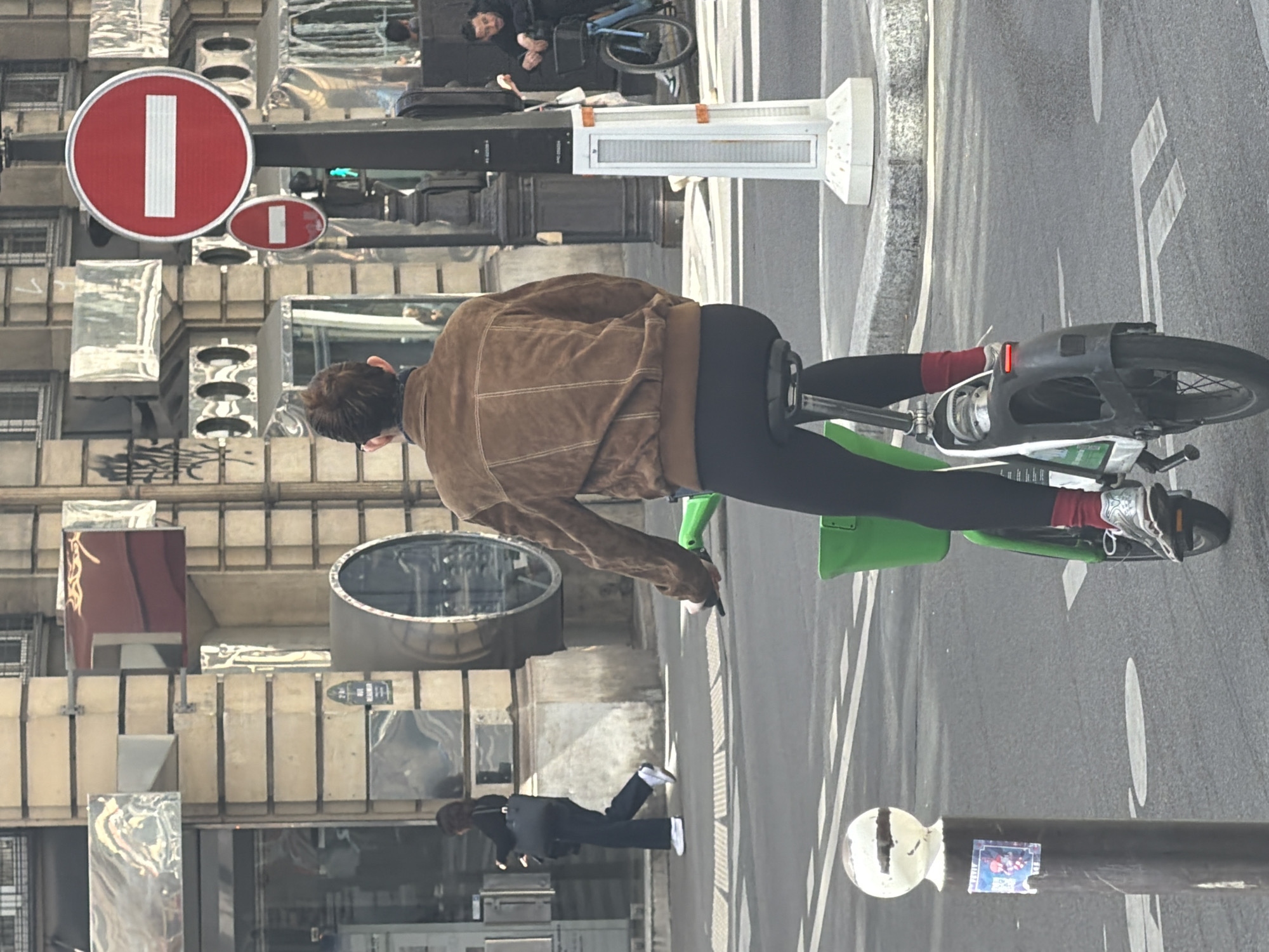 An urban street scene, likely in Paris, France, is depicted during daytime under clear, bright conditions. The central focus is a person riding a bicycle, viewed from behind as they move across the frame. The cyclist wears a brown jacket, black pants, red socks, and white sneakers, and is on a bicycle with a distinctive green frame and dark wheels.
To the left of the cyclist, multi-story buildings line the street, featuring windows, reflective surfaces, and varied architectural elements. Two prominent circular red "No Entry" signs, each with a white vertical bar, are mounted on a structure associated with these buildings. A green traffic light is also visible in the background, suggesting active traffic management.
The street surface on the right displays white road markings on the asphalt. A street pole with a spherical white top and a small, unidentifiable sticker is in the lower foreground. Another person walks on the sidewalk to the left, dressed in dark attire with white shoes and carrying a bag. Further in the background, other individuals are visible, including one sitting on a bicycle and another standing near a building. The overall impression is one of a busy city environment.
