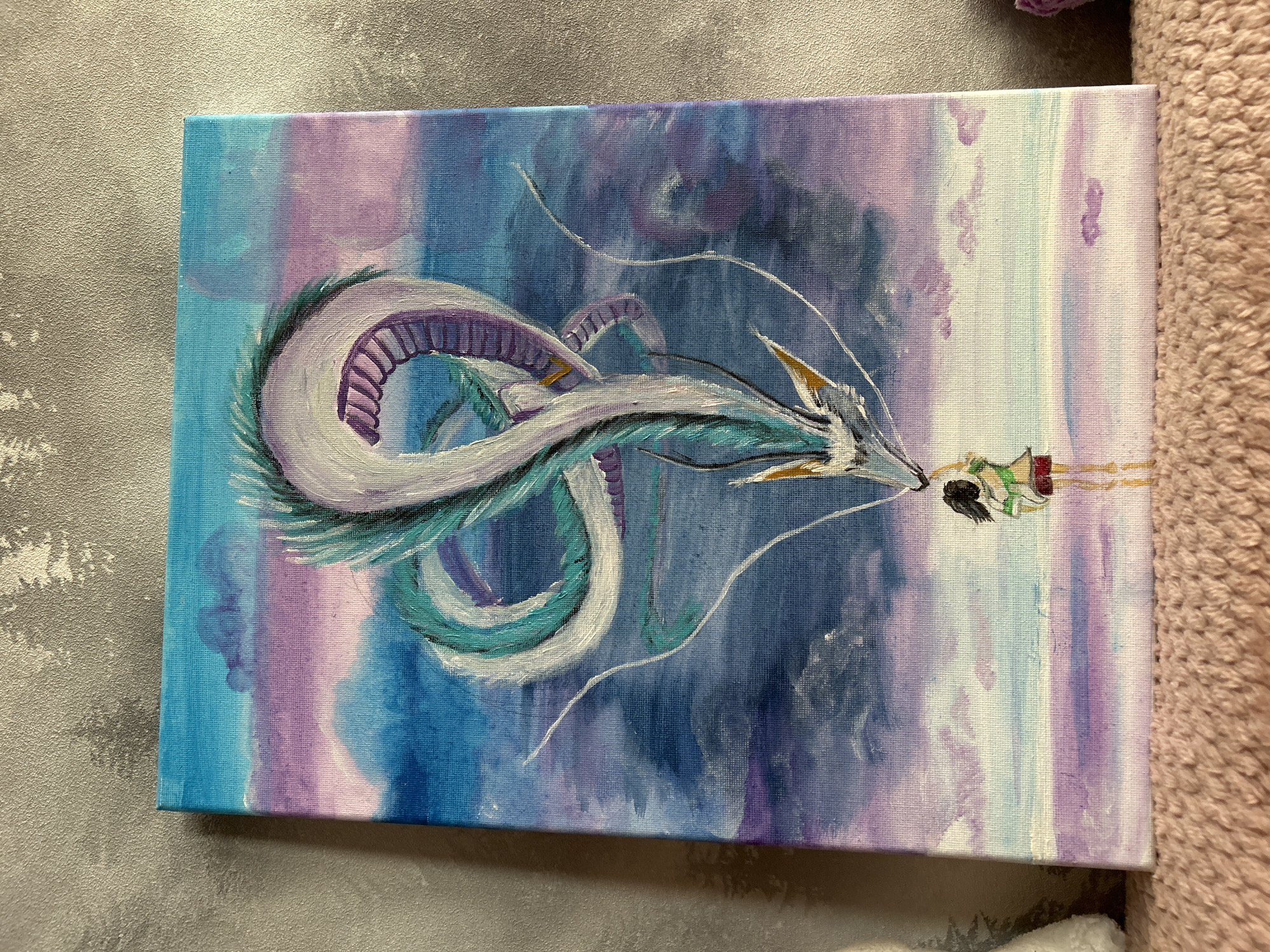 A vibrant acrylic painting on canvas depicts a scene from the animated film "Spirited Away." The central subject is Haku, the dragon spirit, rendered in flowing white, turquoise, and purple hues, with prominent dorsal fins and a long, serpentine body that curves across the canvas. His head, adorned with golden accents, is turned towards a small human figure. This figure, presumably Chihiro, is painted in a simplified style with green and red clothing, holding a small red object towards the dragon.
The background is an abstract blend of watercolor-like washes in shades of blue, purple, and pink, suggesting a dreamlike or ethereal setting. The sky transitions from a deep blue on the left to lighter purples and pinks on the right, with vertical streaks of white and pale blue. The overall mood is mystical and serene, with the gentle interaction between the dragon and the child at its core. The painting is laid flat, with a textured grey surface visible to the left and a soft, fuzzy pink fabric to the right, suggesting it may be displayed or photographed in a home setting. No visible text is present in the image.