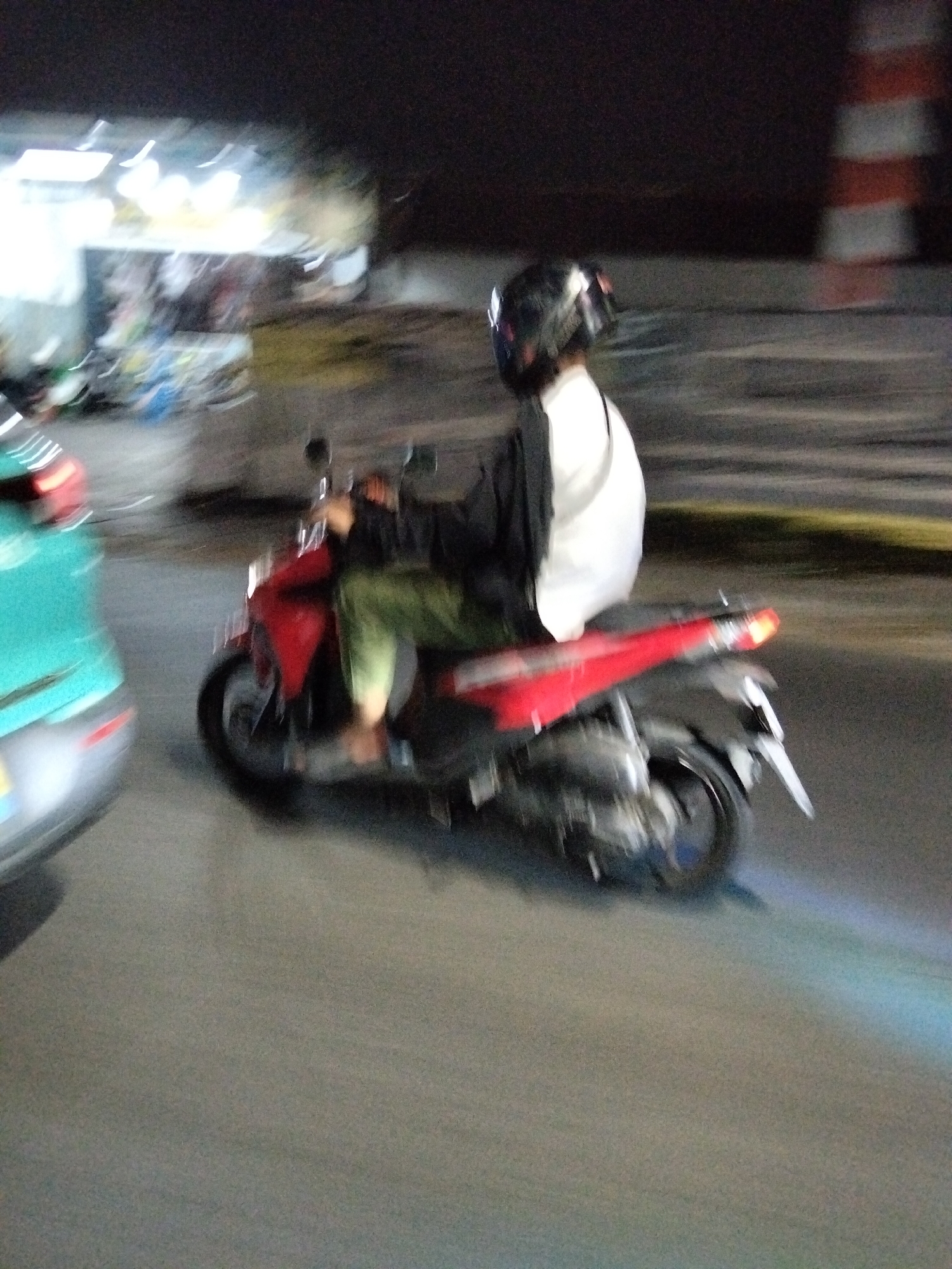 The image depicts a motion-blurred scene at night, likely on a city street in South Tangerang, Indonesia. A person wearing a black helmet and a white shirt is riding a red motorcycle. The rider is also wearing green pants. In the background, there is a blurred view of street lights and possibly a building or stalls. To the left, a portion of a teal-colored vehicle is visible, also motion-blurred. The overall impression is of movement and the nocturnal urban environment.