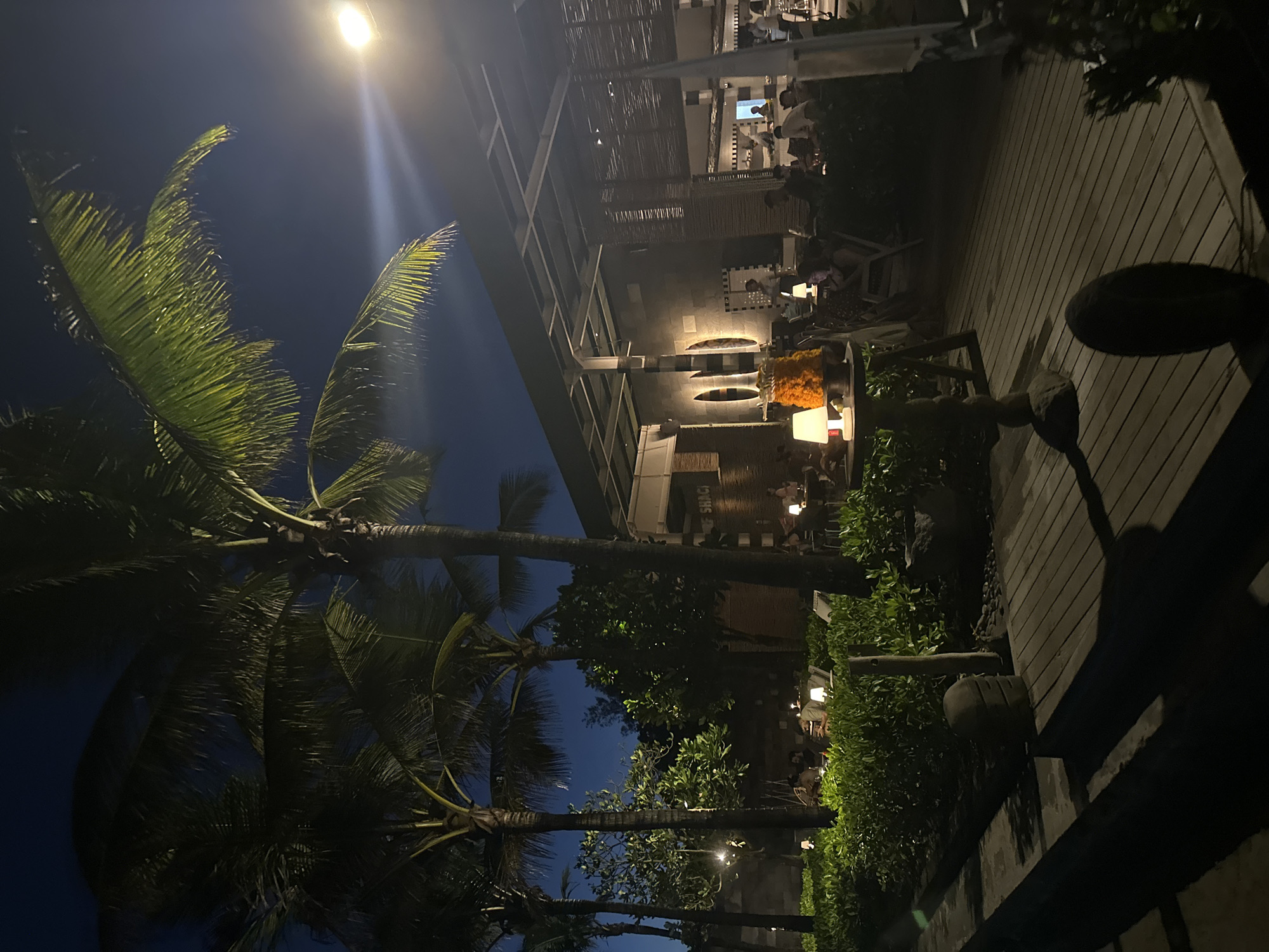 The image captures a vibrant outdoor evening scene at a tropical restaurant or resort in North Kuta, Indonesia. The photograph is oriented vertically, showing a dark, clear night sky with significant artificial illumination.
In the foreground and extending along the left side, several tall palm trees dominate, their fronds detailed against the dim sky, especially where illuminated by a bright overhead street light positioned high on the left. The light casts a glow, highlighting the green texture of the palm leaves and creating lens flares.
On the right side of the frame, a spacious wooden deck with parallel planks is visible, extending towards the viewer. Along the edge of the deck and further into the midground are lush green bushes and smaller tropical trees, forming natural dividers. Scattered within this greenery and on patios are multiple outdoor tables where people are seated. Each table is illuminated by small, modern table lamps, casting a warm glow. One table features a decorative arrangement of orange-yellow flowers.
Further back, a multi-level building with an open-air design appears to be the main restaurant or bar structure. It features a woven or slatted ceiling, dark wall panels, and structural elements. More patrons are visible dining or socializing at tables both within the building's open sections and on an upper-level patio. Wall sconces add to the ambient lighting.
The overall activity depicts people enjoying an evening meal or drinks in a relaxed, social atmosphere. The dark sky, combined with the extensive use of warm artificial lighting, sets a sophisticated nighttime ambiance. Part of a sign on the building can be seen, reading "SHACK."