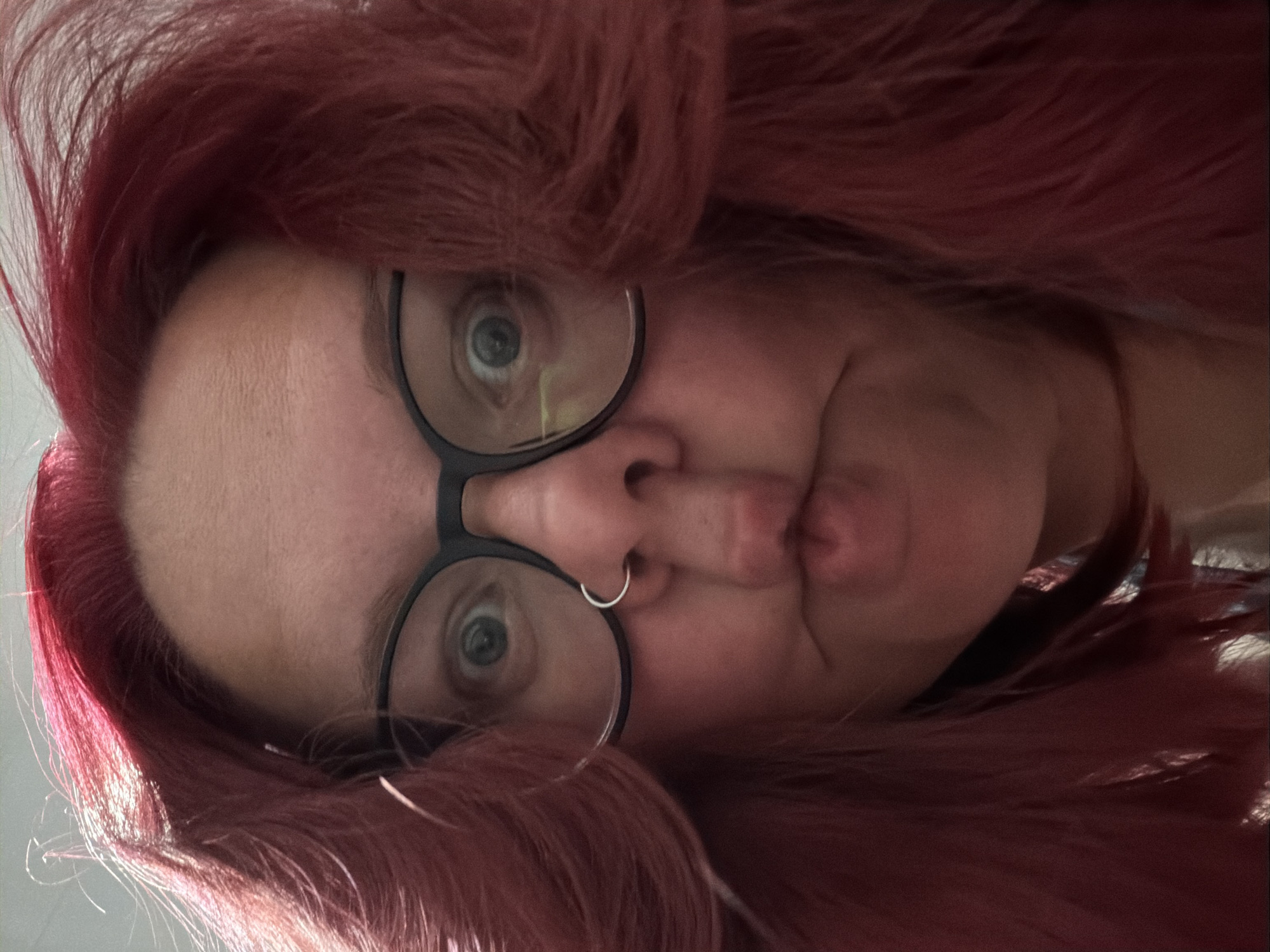 A close-up, low-angle portrait captures an adult individual with vibrant red, wavy hair that frames their face. They wear black-framed, round eyeglasses and a small silver hoop piercing in their right nostril. The individual's eyes are wide open, looking directly at the viewer, and their lips are prominently puckered in an exaggerated "fish face" expression. Fine lines are visible on their forehead. The background is indistinct and uniformly light, suggesting an indoor environment without specific identifiable features. The image provides no visual indicators to confirm a specific geographic location such as the city of Chaikovsky, Russia.