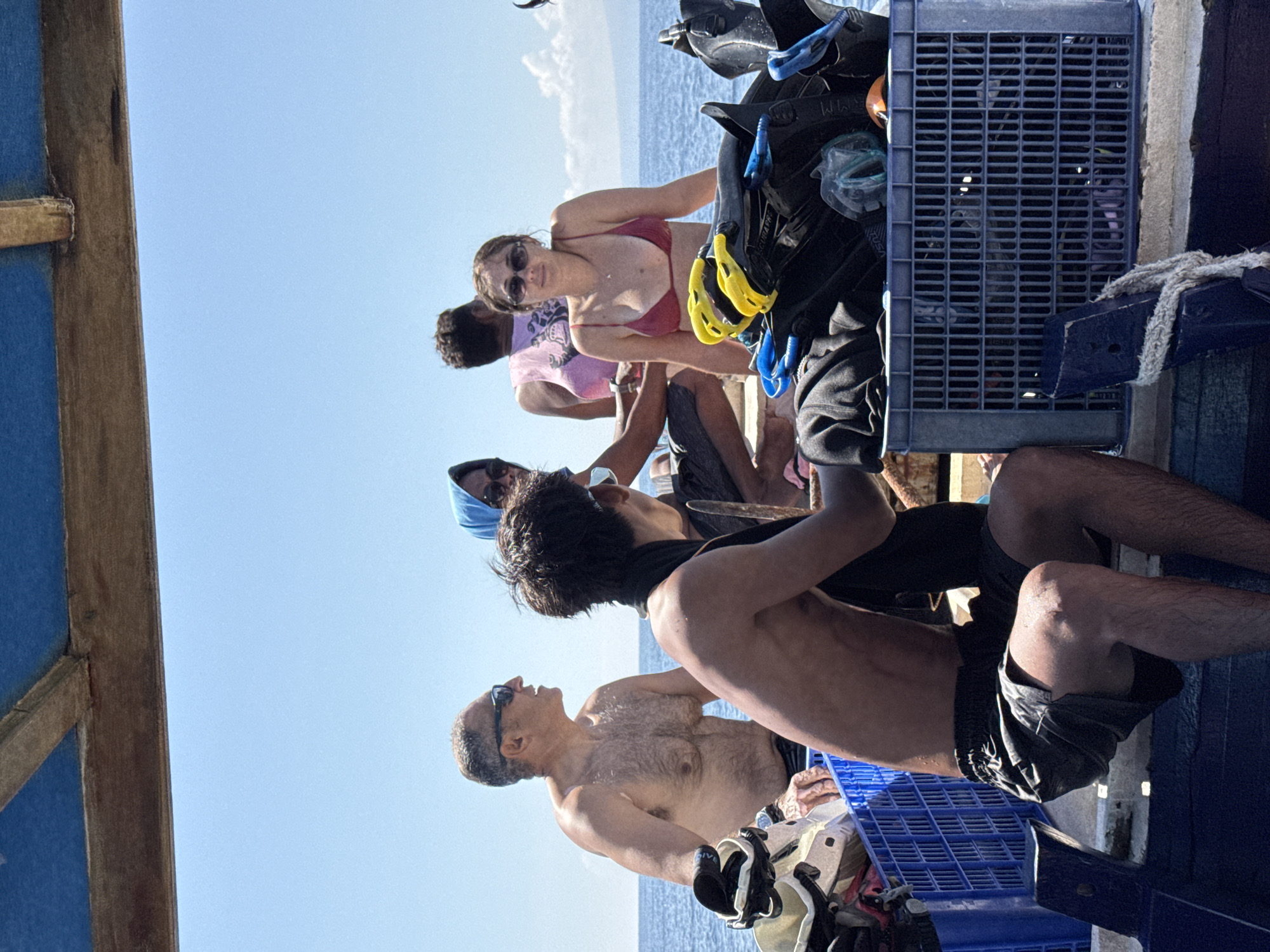 The image shows a group of people preparing for a dive, possibly on a boat off the coast of Indonesia. Visible are divers in swimwear and dive gear. There are blue plastic crates holding diving equipment. The location is the city of Unknown, Indonesia. The environment includes the sky and ocean.