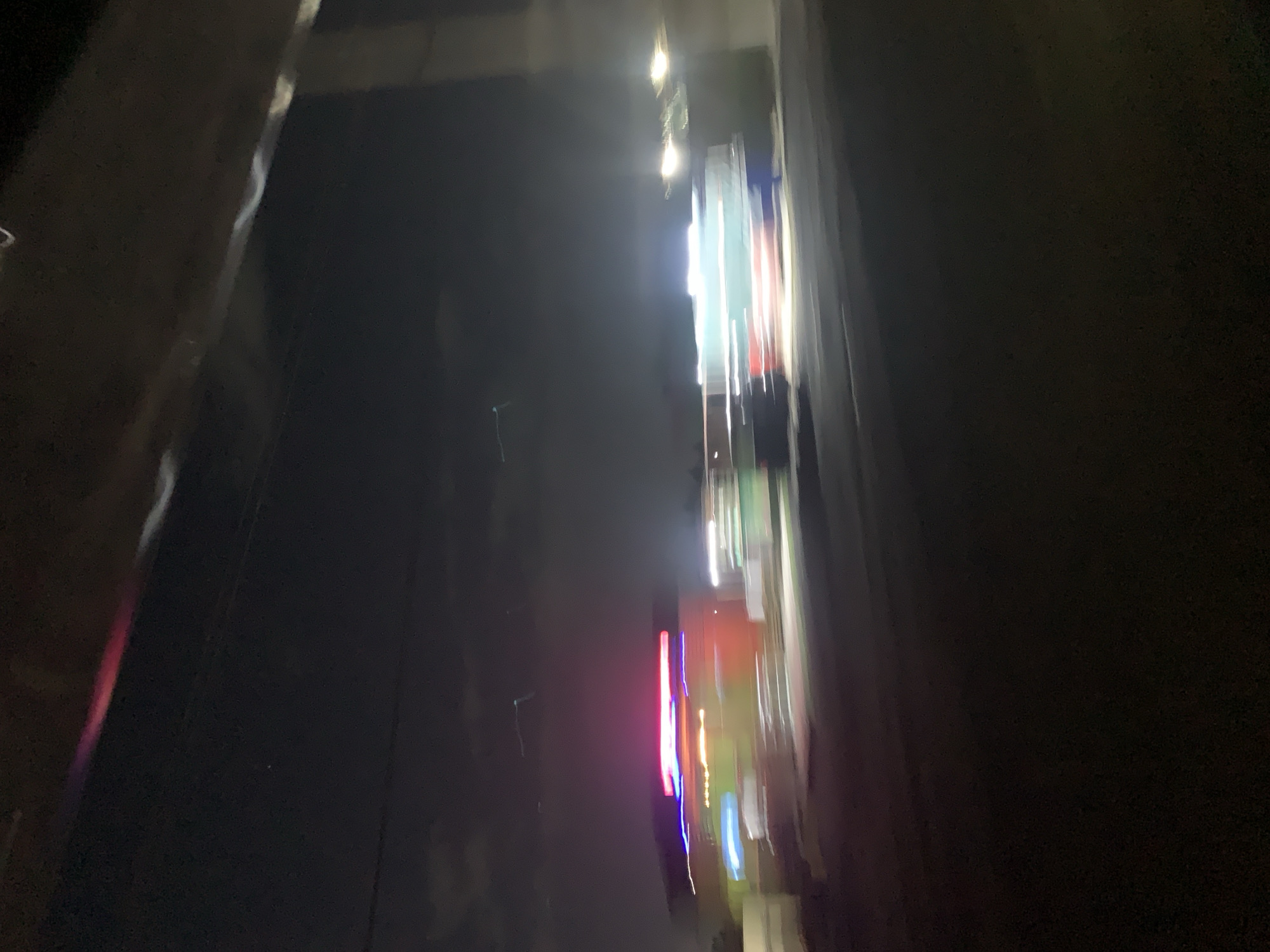 The image shows a blurred view of a cityscape at night. Bright lights from buildings and signs are visible as streaks of color, predominantly white, green, red, and blue. The surrounding environment appears dark, suggesting a nighttime setting. Based on the prompt, the location is assumed to be in Dar es Salaam, Tanzania, although there are no identifiable landmarks to confirm this from the image. Due to the motion blur and low light conditions, no people, specific objects, or actions are clearly discernible. The focus is on the lights of the city and the resulting streaks caused by movement during the image capture.