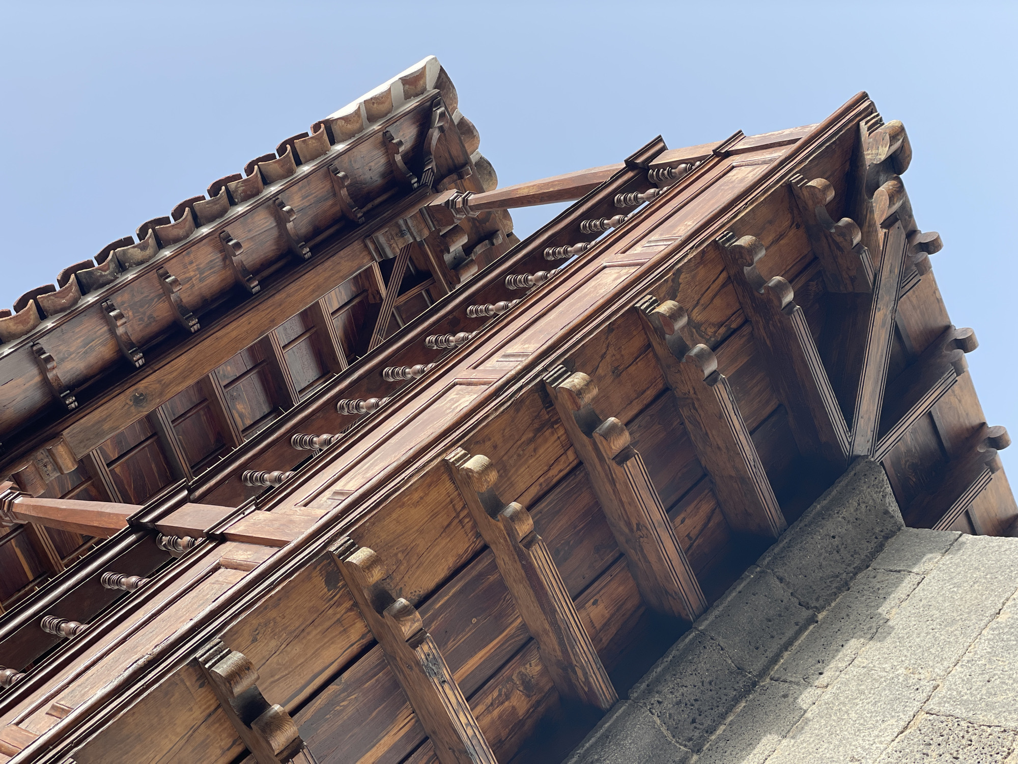 This image depicts the eaves and roof of a traditional Canarian building in Santa Cruz de Tenerife, Spain. The architecture features intricately carved dark wood beams, corbels, and a detailed eave design. A section of the roof is tiled with terracotta-colored barrel tiles. The eaves overhang a gray stone wall. The sky is a clear, light blue. The perspective is a low-angle shot, looking upward at the building's structure.