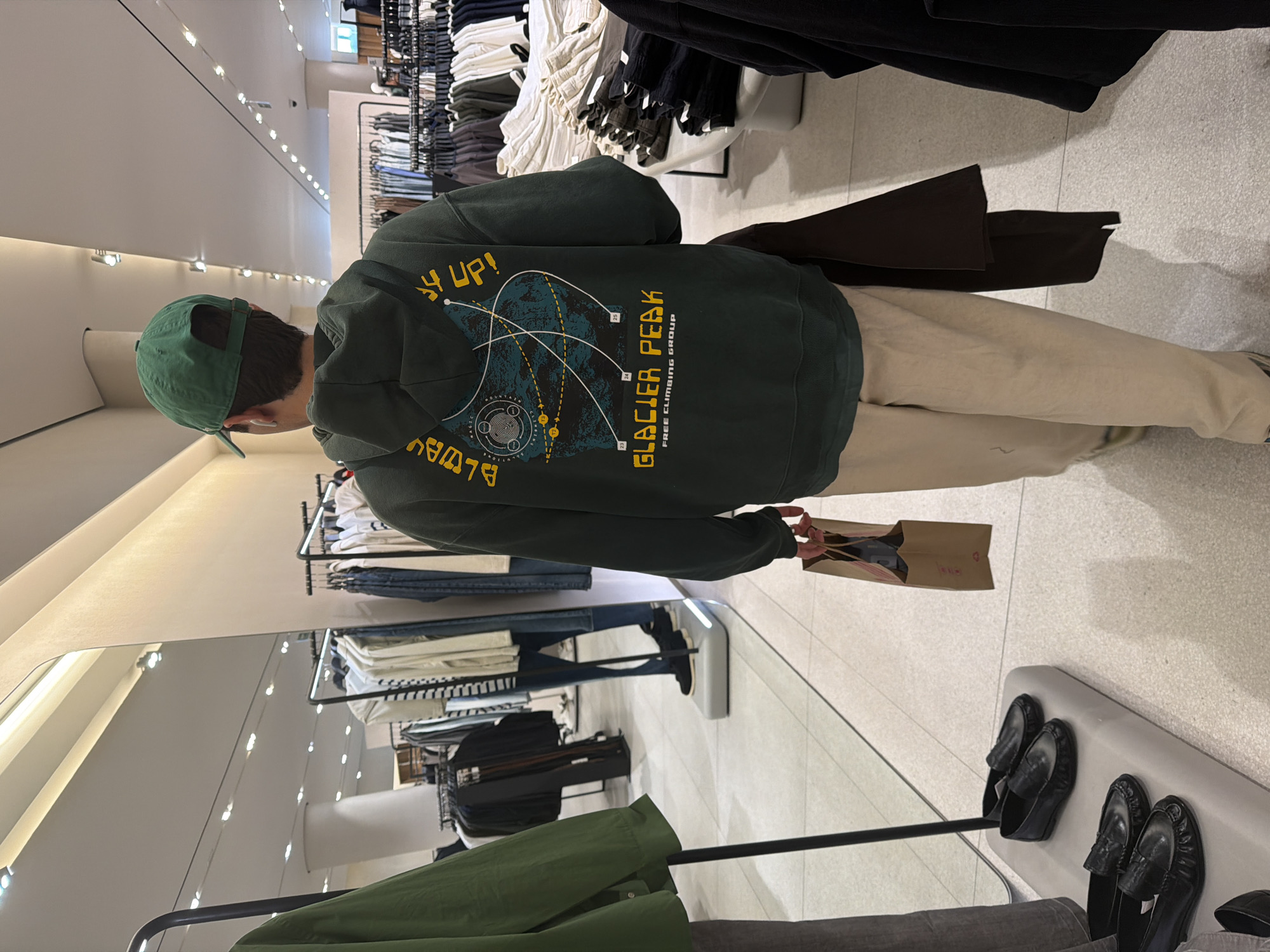 A person with a green cap stands in a clothing store with clothing racks and mirrors. The person is wearing a dark green hoodie with "Glacier Peak Free Climbing Group" written on the back and is holding a brown shopping bag. Several pairs of black shoes are displayed on a white shelf. The location is not specified and cannot be identified as Puebla, Mexico based solely on the given image.