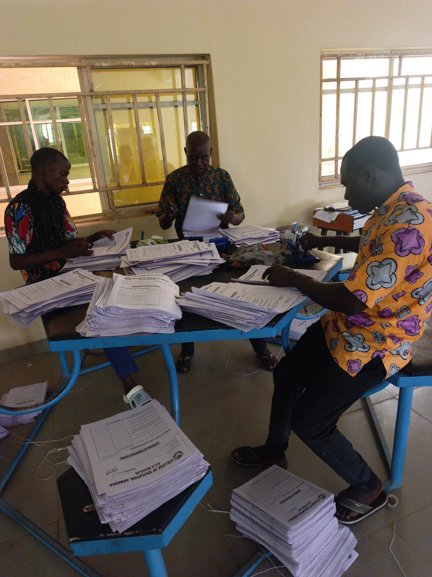 Three adult men are shown indoors, seated at light blue-framed tables with dark tops, busily engaged in sorting and processing numerous stacks of papers. The room has plain, light-colored walls and multiple windows with metal grates, allowing bright daylight to illuminate the scene, suggesting it is daytime.
On the left, a man wearing a black shirt with vibrant, colorful patterns is handling a pile of documents. In the background, a third man with a patterned shirt is also focused on paperwork. To the right, a man in a bright orange shirt with distinct purple and gray patterns is actively using a stamper on documents stacked in front of him. Large quantities of white papers are distributed across the tables and even on small stools below, indicating a significant administrative task.
Visible text on the documents clearly identifies them with "COLLEGE OF EDUCATION, AKWANGA", "N.C.E REGULAR", and "MORNING/AFTERNOON/EVENING", along with "DIRECTIONS FOR CANDIDATES" and "TOTAL (0)". A "STAMP" device is also visible on the table. These details suggest the men are likely involved in administrative work such as processing examination scripts or registration forms for an educational institution. The men appear focused and diligent in their work within what seems to be an office or administrative room.