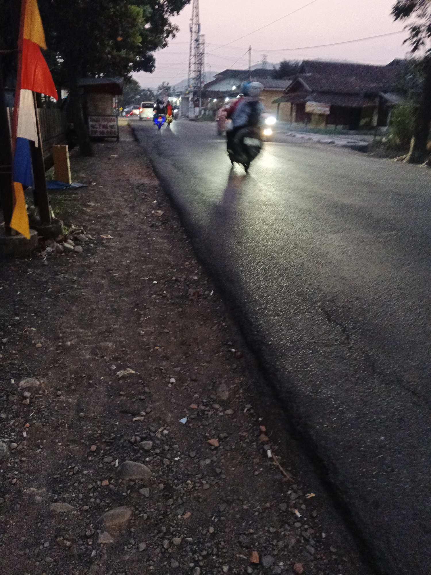The image depicts a roadside scene at dusk in Sukabumi, Indonesia. The foreground is a gravelly dirt path bordering a paved road. Along the left edge of the path, a wooden pole supports several colorful flags, red, white, yellow, and blue, which appear to be decorative or symbolic. Behind the flags, a dark tree with dense foliage stands next to a small roadside stall or kiosk. The road itself is wet and reflects the light from a motorcycle that is the primary subject in motion. The motorcycle, with a rider wearing a blue helmet, is moving away from the camera towards the right side of the frame. The motorcycle's headlight is on and illuminates the wet road. In the background, houses with traditional tiled roofs are visible, suggesting a semi-rural or suburban setting. A tall telecommunications tower is also prominent in the distance, along with other structures. The sky is a pale pink, indicating the twilight hours. The overall impression is a quiet street scene during the transition from day to night.