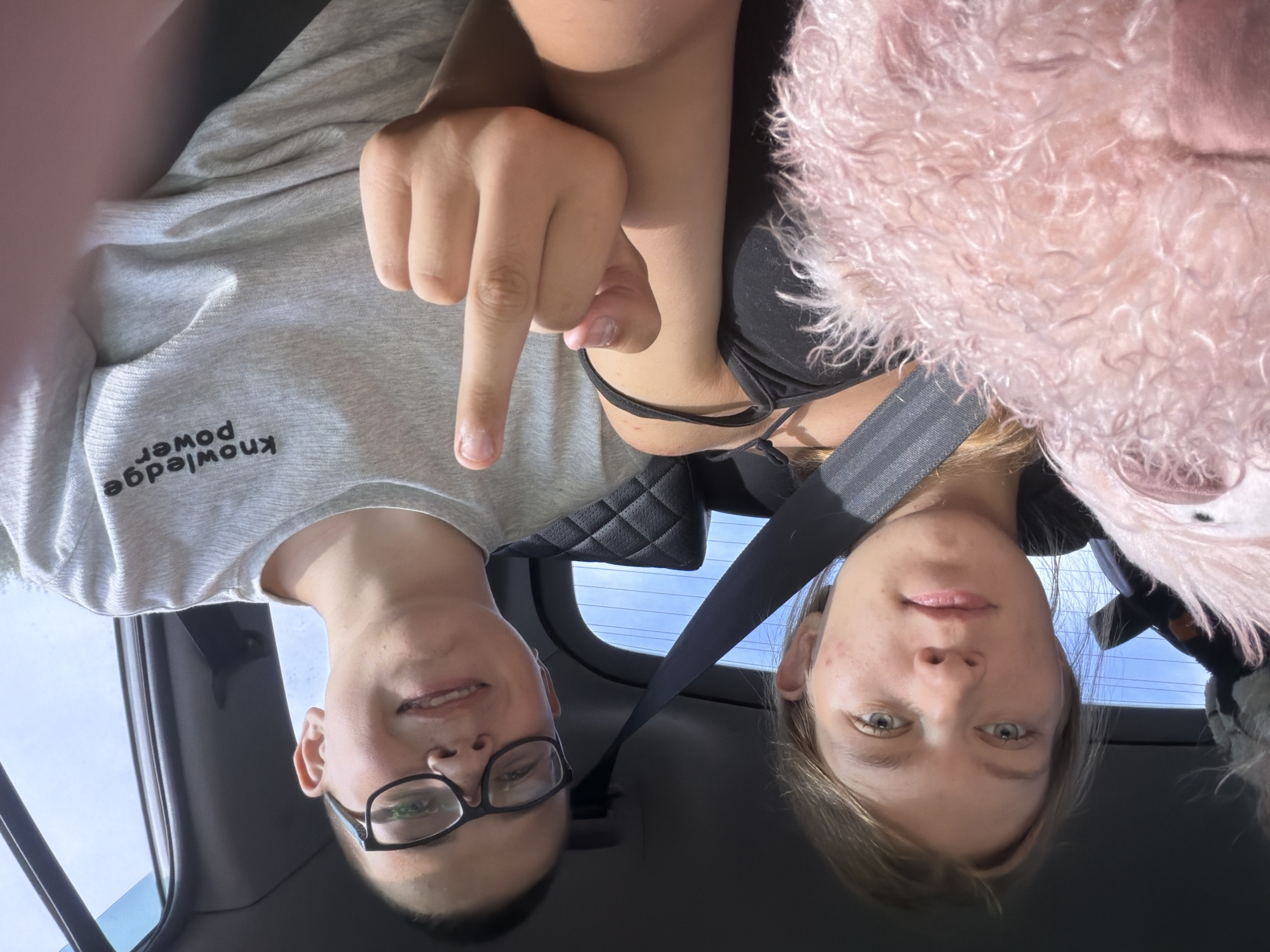 In the image, two young individuals, a female on the left and a male on the right, are seated inside a vehicle. The female has blonde hair and blue eyes, and she is holding a pink plush toy. She is wearing a black top. The male has short dark hair and is wearing glasses and a grey t-shirt with the text "knowledge power" printed on it. He is holding up his middle finger towards the camera. The vehicle appears to be a car, with a seatbelt visible across the female and the interior of the car visible in the background, including a headrest. The lighting suggests it is daytime, with sunlight filtering through the windows. The location is identified as Novoye Mavrino, Russia.