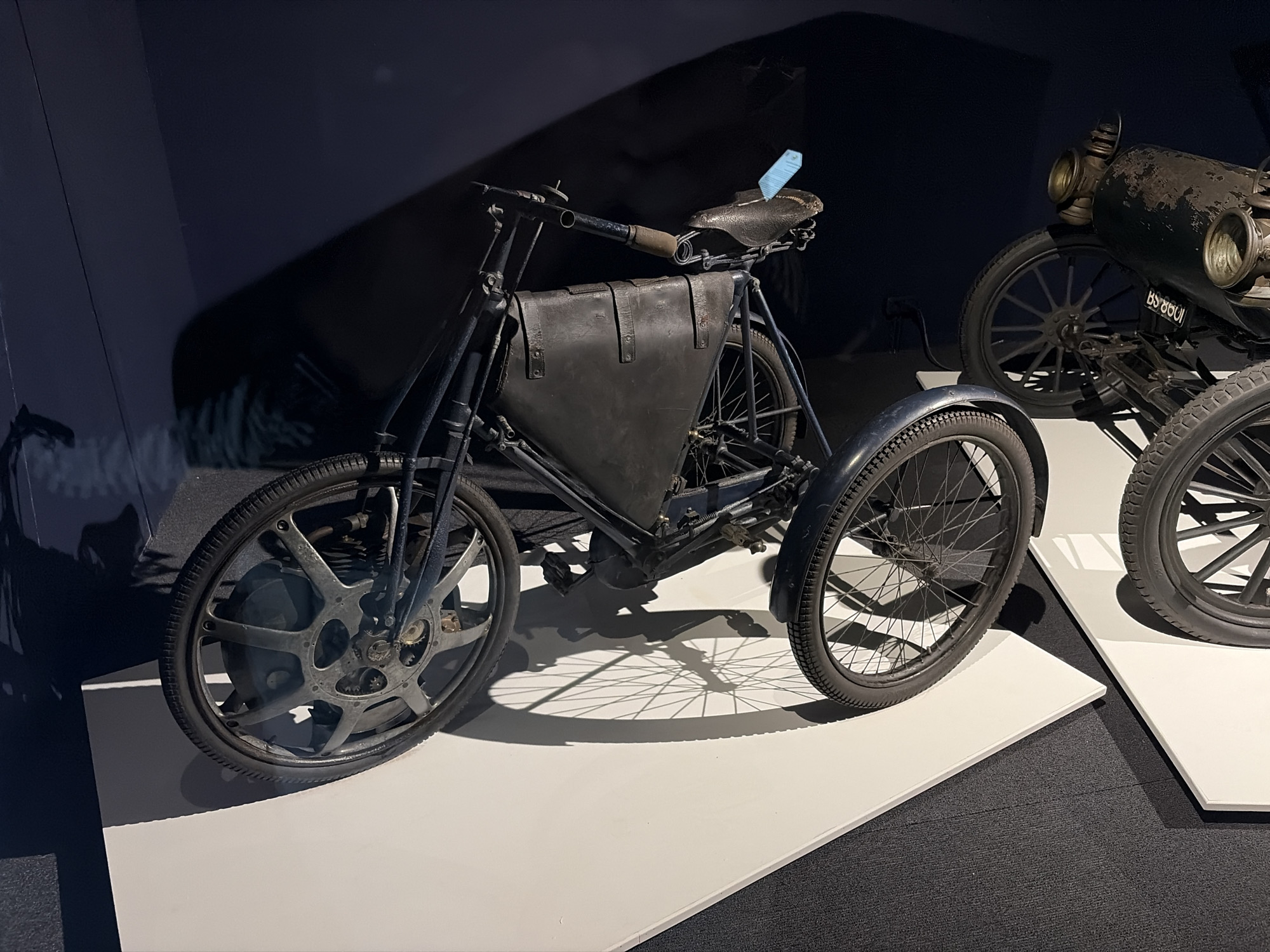 An antique, three-wheeled vehicle, likely an early motor tricycle, is prominently displayed in what appears to be a museum or exhibition setting. The vehicle is predominantly dark-colored, featuring a robust frame, large spoked wheels with rubber tires, and a worn leather-like saddle. A substantial, dark, possibly leather, storage bag or compartment is secured within the frame, positioned between the front wheel and the seat. The front wheel stands out with its intricate design, resembling a solid disc with circular cutouts that reveal internal mechanical components, such as gears. A fender is visible over the right rear wheel.
In the background, another antique vehicle is partially visible to the right. This vehicle, possibly an early car or motorcycle, features a large, dark, cylindrical structure (perhaps an engine or fuel tank) and distinctive headlights. Its dark spoked wheels are also visible, and a license plate clearly shows the text "BS 8801".
Both vehicles are stationary and presented on clean, white display platforms that contrast sharply with the dark, carpeted floor of the exhibition space. The setting is dimly lit, with spotlights creating dramatic lighting effects and casting distinct shadows of the vehicles onto the platforms. A small, light blue tag is attached to the saddle of the tricycle, likely an informational label for visitors. No people are present, and the scene depicts a static display of historical transportation artifacts.