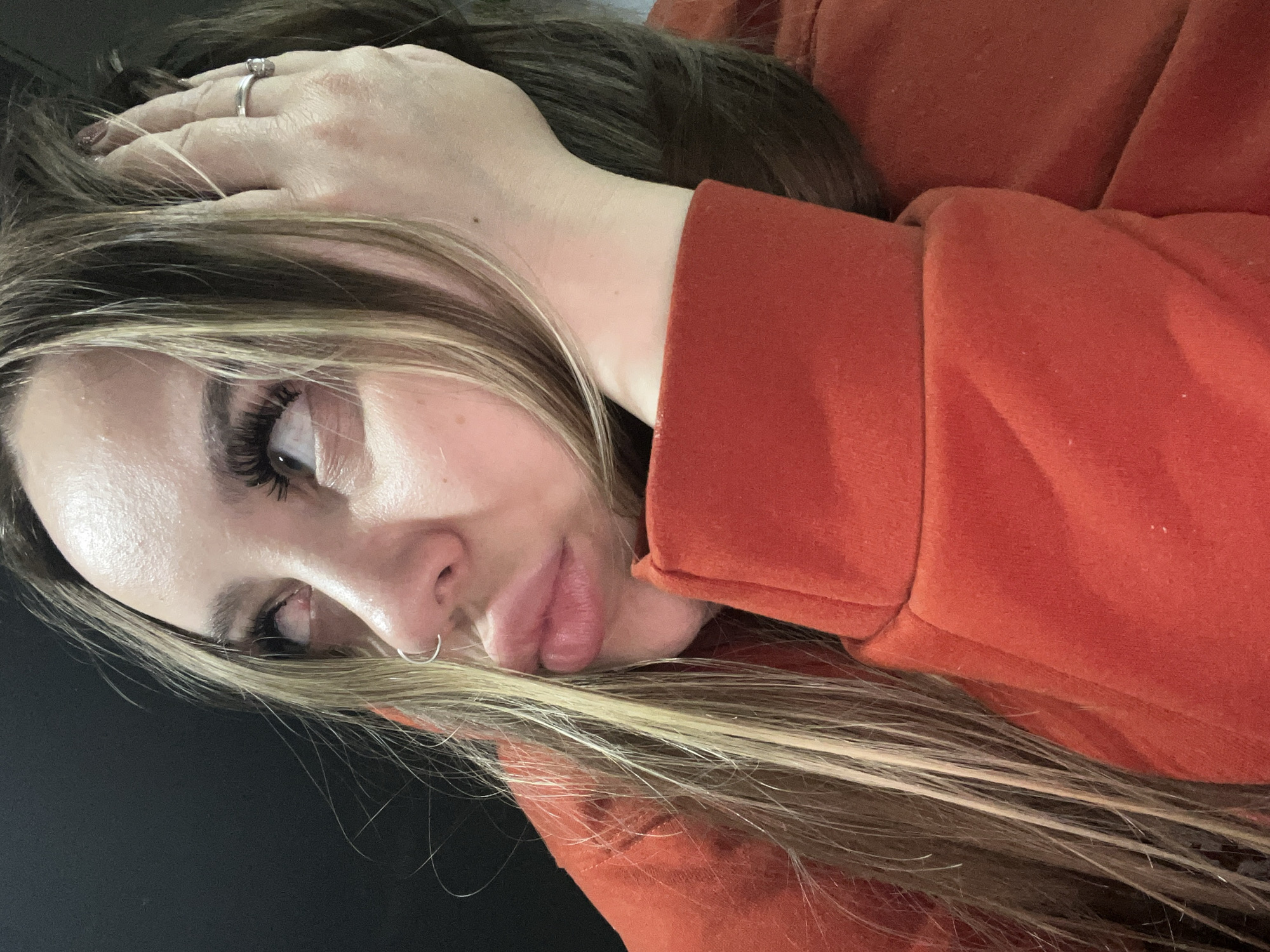 A woman is lying down, wearing an orange sweatshirt. Her head rests on her hand, and her hair is a mix of brown and blonde. She has a nose ring and is looking off to the side. The background appears to be a dark, possibly black, surface. There is no information provided in the image to determine if the scene is in Krasnodar, Russia, or anywhere else. The image provides no details to suggest the location.