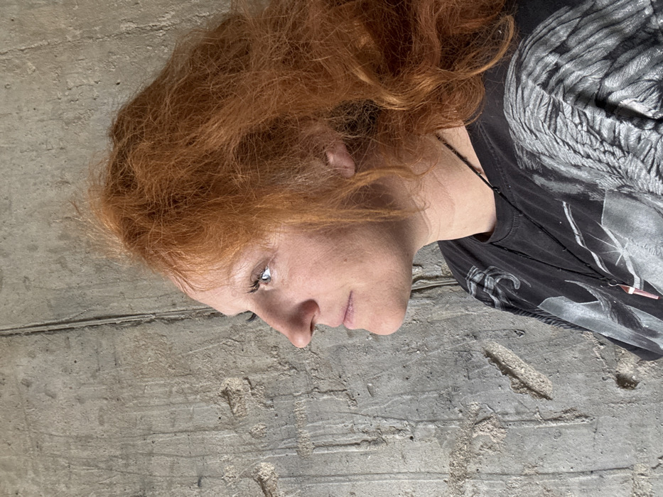 An adult female is depicted in a left-facing profile against a textured concrete wall. She has voluminous, reddish-orange, wavy hair and a fair complexion. Her left eye is open, and she is looking slightly upward and forward. She wears a dark t-shirt with a grey, possibly avian or feathered, graphic print. A thin black necklace with a light-colored, rectangular pendant is visible around her neck. The concrete wall behind her is rough, with vertical lines and irregular surface variations. The specific location within the city of Chaikovsky, Russia, cannot be determined from the image.