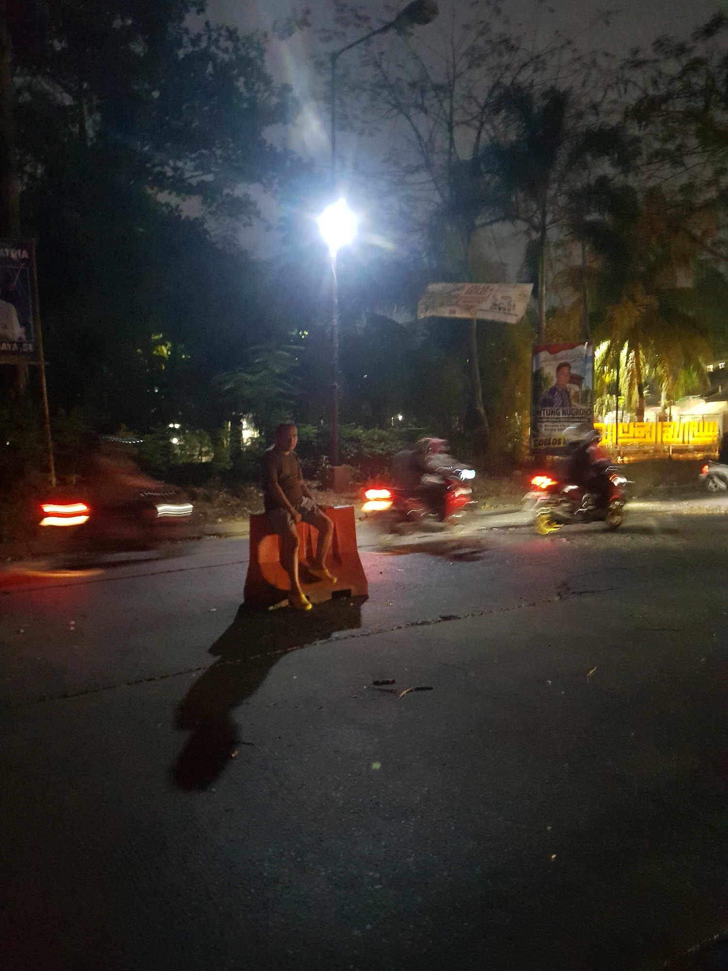 In Karangsatria, Indonesia, at night, a person sits on an orange traffic barrier on a paved road. The individual, wearing a dark t-shirt and shorts, is facing the camera with their legs crossed. The road is wet and reflects the light from a streetlamp and passing vehicles. To the left, a car's taillights glow red, creating streaks due to motion blur. Further down the road, two motorcycles are visible, also with motion blur, their headlights and taillights illuminated. In the background, trees and vegetation are dimly lit by the streetlights. Several posters are visible, one on the left with a portrait and text, and another in the center-right, also featuring a portrait. The scene suggests a public street at night, possibly near a road construction or event area due to the traffic barrier.