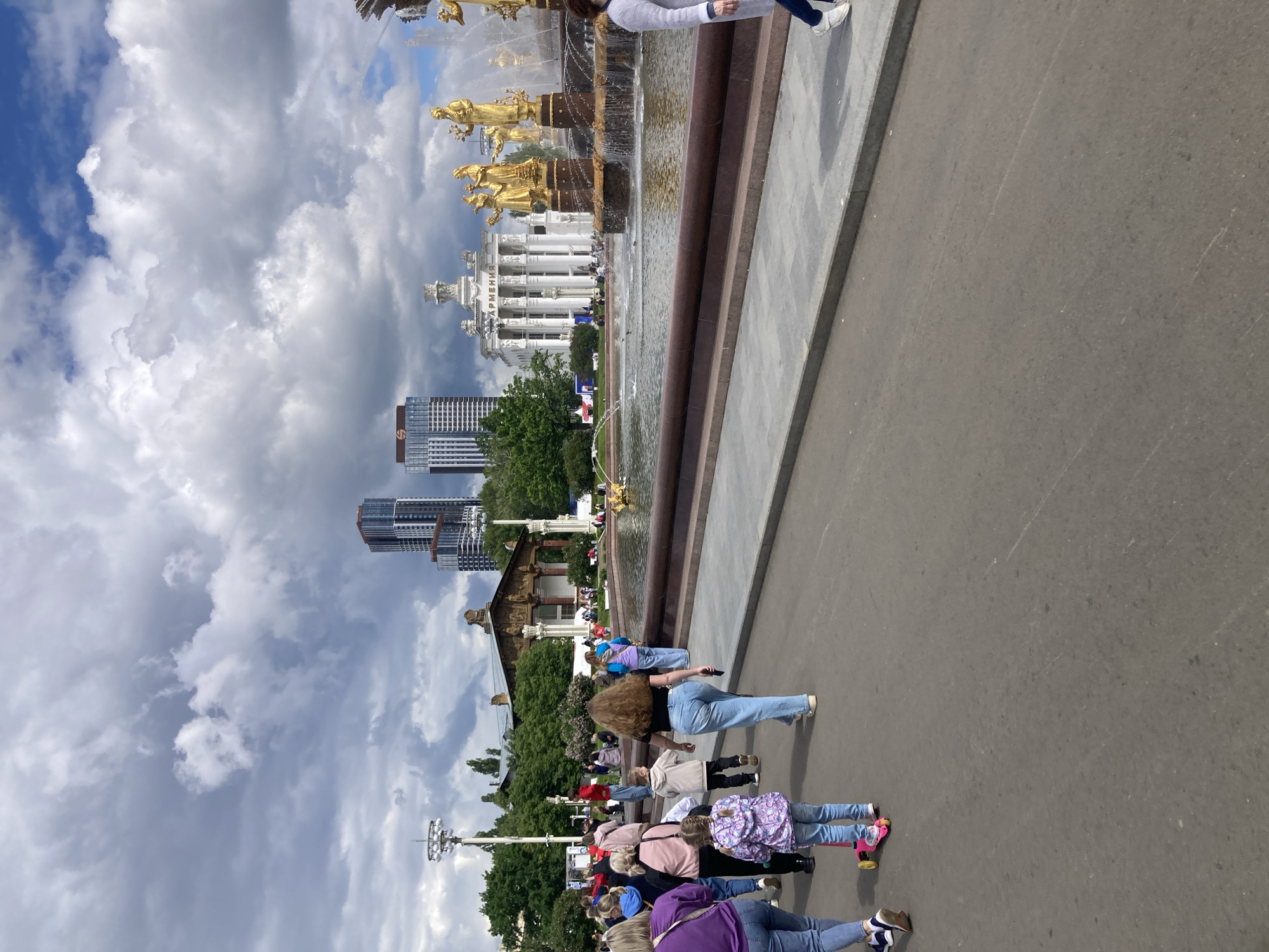 The image shows a view of the Friendship of Nations Fountain in Moscow, Russia. A group of people is walking along a path next to the fountain. The fountain is decorated with golden statues and surrounded by water. Buildings and trees are visible in the background. The sky is cloudy.