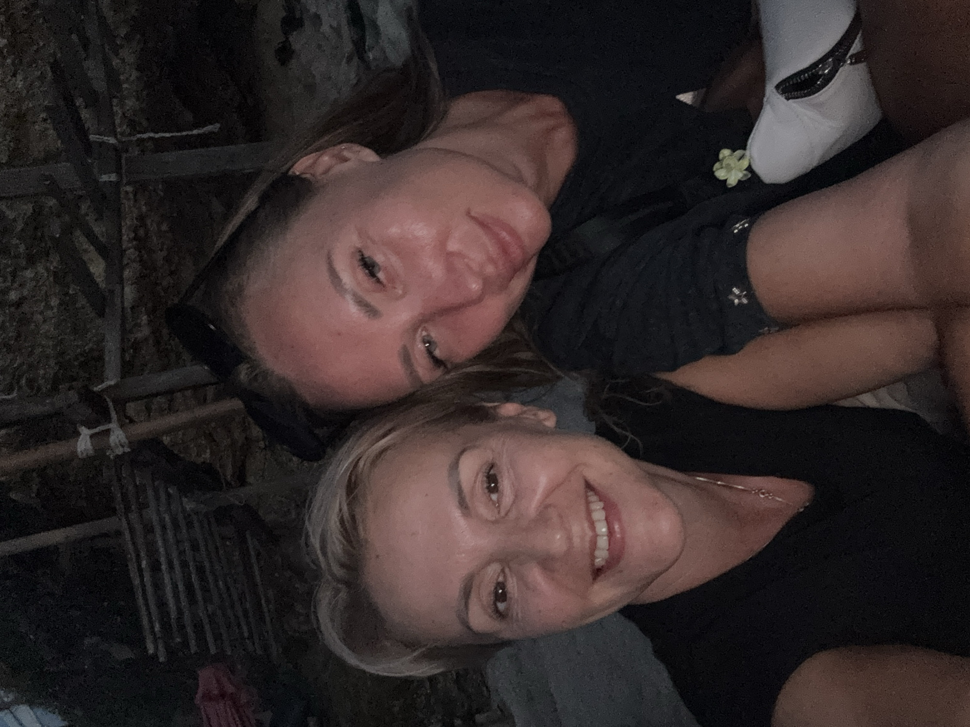 Two women are posing closely together in a selfie. One has long hair and is wearing a black top. The other has shorter, blonde hair and is also wearing a black top. A white sock with a zipper detail is visible, along with what appears to be a small, light-colored flower. The background is dark and includes some sort of wooden or bamboo structure. The location is Kuta Selatan, Indonesia.