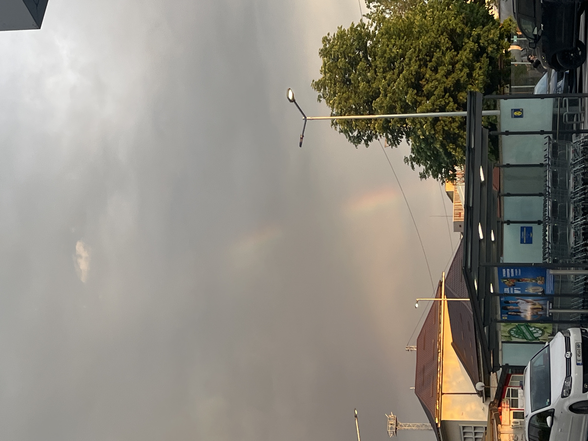 The image, taken in Blagoevgrad, Bulgaria, shows a cloudy sky with a faint rainbow. A streetlight is visible near a tree, while a structure, possibly a bus stop or shopping area, is to the right. A white car is parked in front of the structure. The sky is overcast, and the light suggests it may be late afternoon or early evening.