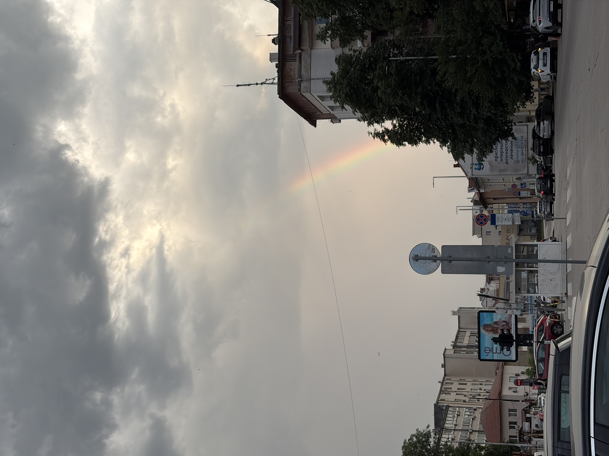 The image depicts a city street scene in Pleven, Bulgaria, under a dramatic sky with a visible rainbow. The sky is filled with a mix of dark, ominous clouds and brighter, illuminated areas, suggesting a recent or ongoing rain shower. A prominent rainbow arcs across the sky, its colors fading into the hazy atmosphere.
In the foreground, a street intersection is visible, with cars driving and parked. The road surface is asphalt. Several buildings line the street, exhibiting varying architectural styles, common in a European city. One building on the right is taller and appears to be an apartment or commercial building, with balconies and an antenna on its roof. Another building on the left has a reddish-brown tiled roof.
A large digital billboard is mounted on a pole in the middle ground, displaying an advertisement with a woman's image. Various street signs, traffic lights, and utility poles are also present, indicating urban infrastructure. A pedestrian crossing is marked on the road.
In the distance, more buildings of different heights and styles can be seen. Trees with green foliage are scattered along the street and around the buildings, adding a touch of nature to the urban environment. A few birds are visible in the sky, appearing as small dark specks.
The overall impression is a snapshot of city life during or immediately after a weather event, with the striking visual of the rainbow adding a touch of natural beauty to the otherwise mundane urban landscape. The presence of a billboard advertising a product or service, along with the cars and buildings, firmly places the scene in a developed urban area. The Cyrillic script on some of the signs confirms the location as Bulgaria.