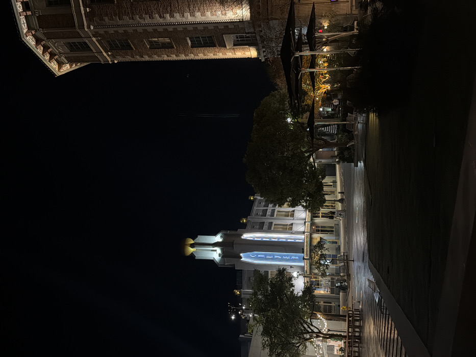 A night scene features two contrasting architectural styles. At the top, a brick building with ornate details is partially visible against the dark sky. Below, a modern building with illuminated features, including a blue-lit vertical element, stands prominently. Trees frame the modern structure, and a paved area with seating and sparse lighting suggests a public space. The composition is shot from a low angle, accentuating the height and scale of the buildings.